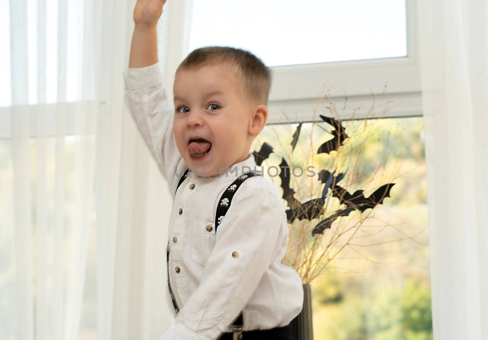 Halloween concept. Autumn holiday. A boy dances with his tongue hanging out against the background of a window near a black vase with bats cut out of paper on dry branches. Close-up. Background.