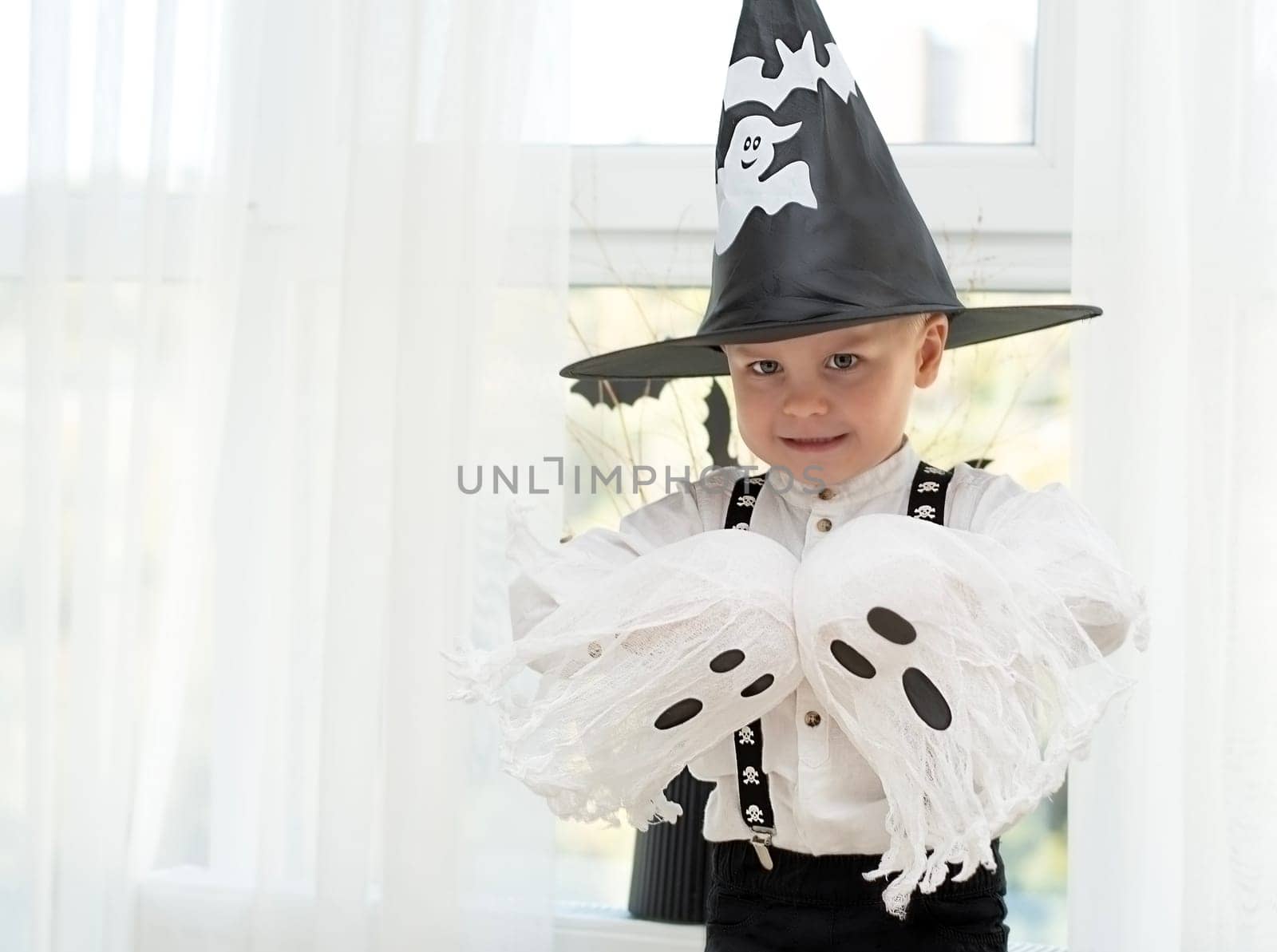 Halloween concept. A small handsome boy in a wizard's hat with white ghosts plays cheerfully and emotionally in a home interior against the background of a window. Close-up.
