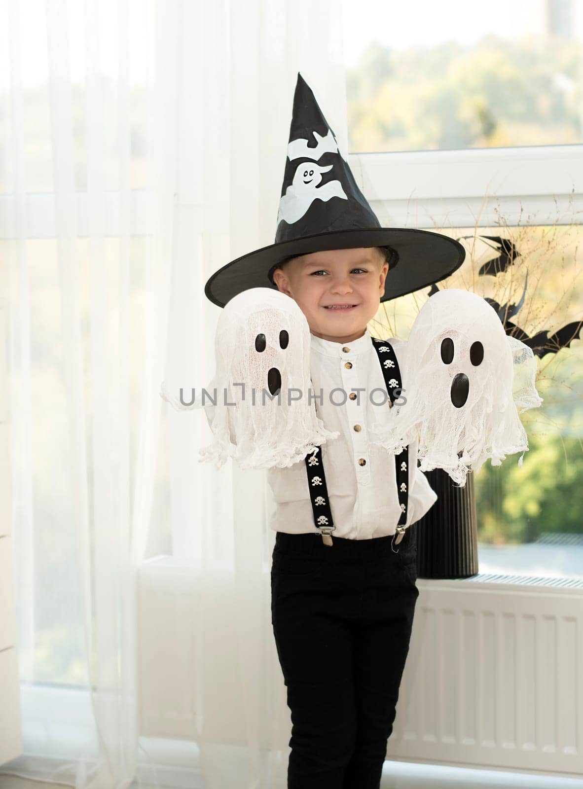 Halloween concept. A small handsome boy in a wizard's hat with white ghosts plays cheerfully and emotionally in a home interior against the background of a window. Close-up.