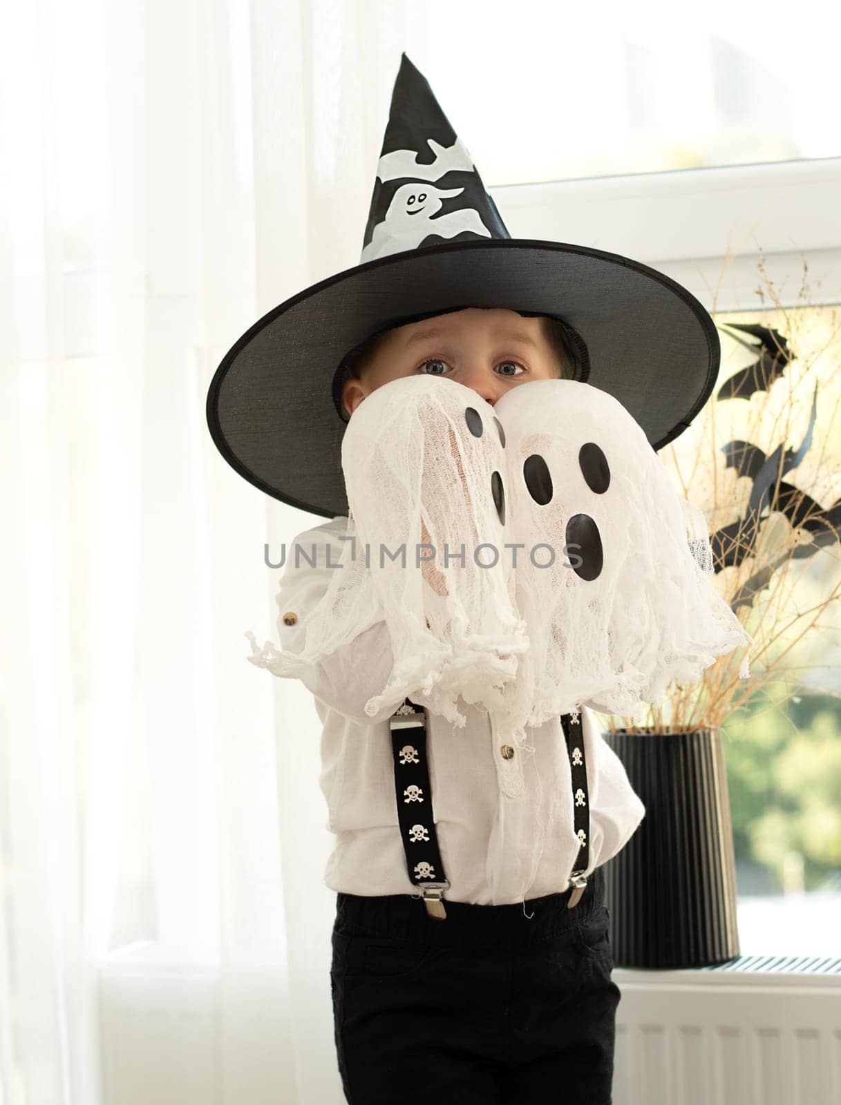 Halloween concept. A small handsome boy in a wizard's hat with white ghosts plays cheerfully and emotionally in a home interior against the background of a window. Close-up.