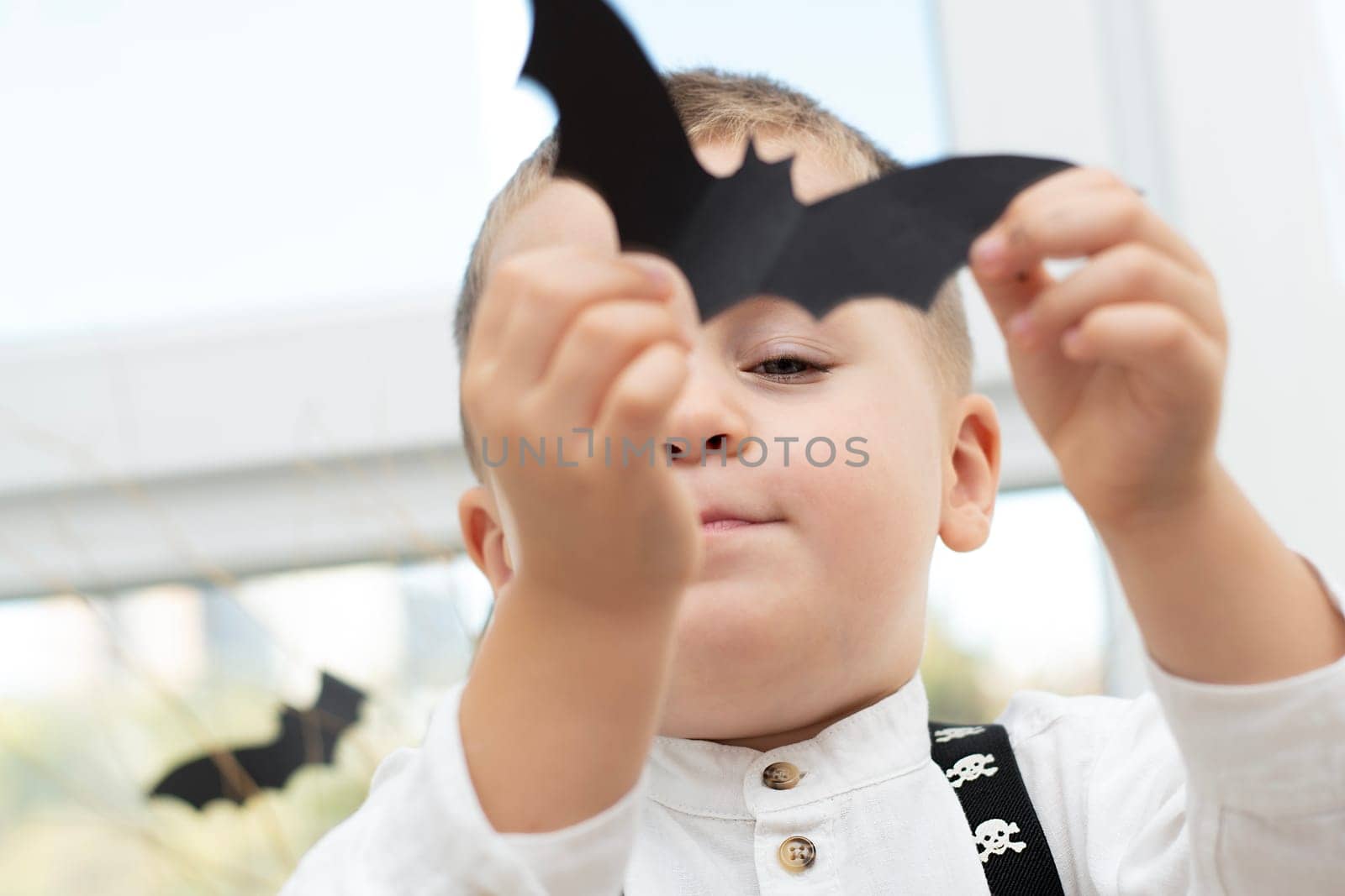 Halloween concept. A small handsome boy emotionally holds a bat made of black paper in his hand. Portrait. Close-up. Soft focus.