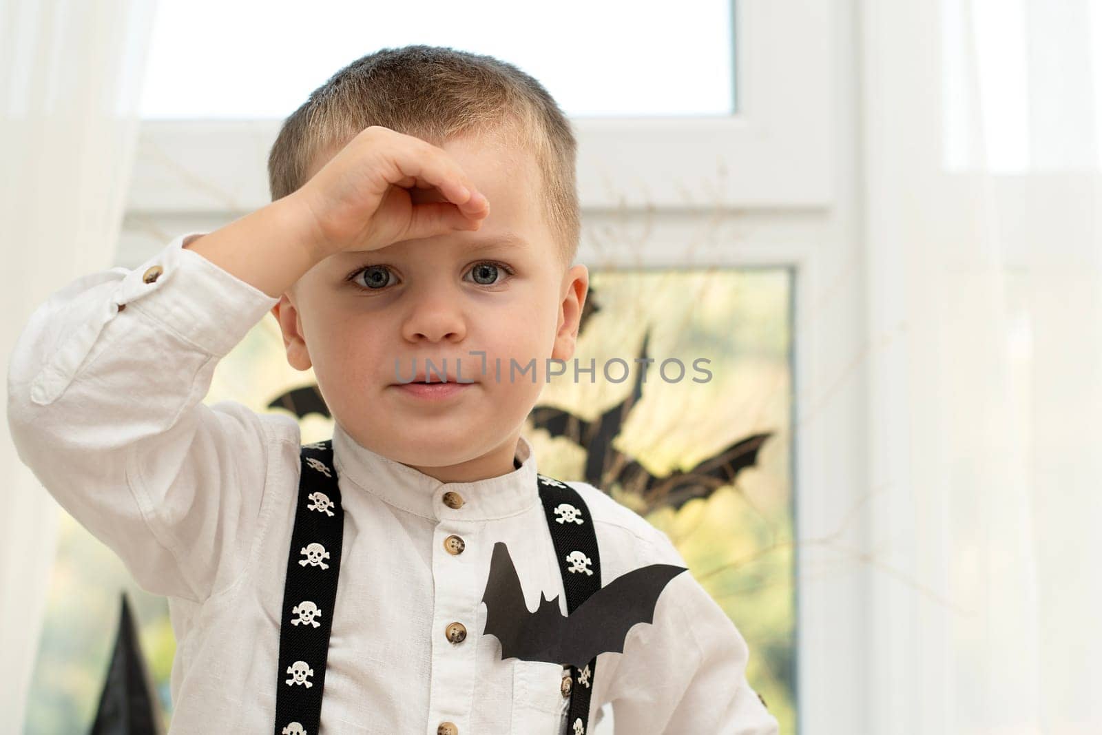 Halloween concept.A handsome boy,with emotional facial expressions, in suspenders with skulls against the background of a window with a vase with dry branches and black paper bats.Close-up.Soft focus.