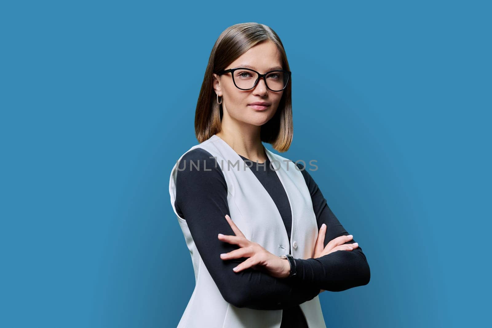 Serious confident young woman with eyeglasses, crossed arms posing on blue studio background. Attractive female in black and white clothes. Beauty fashion style youth business job work staff employee