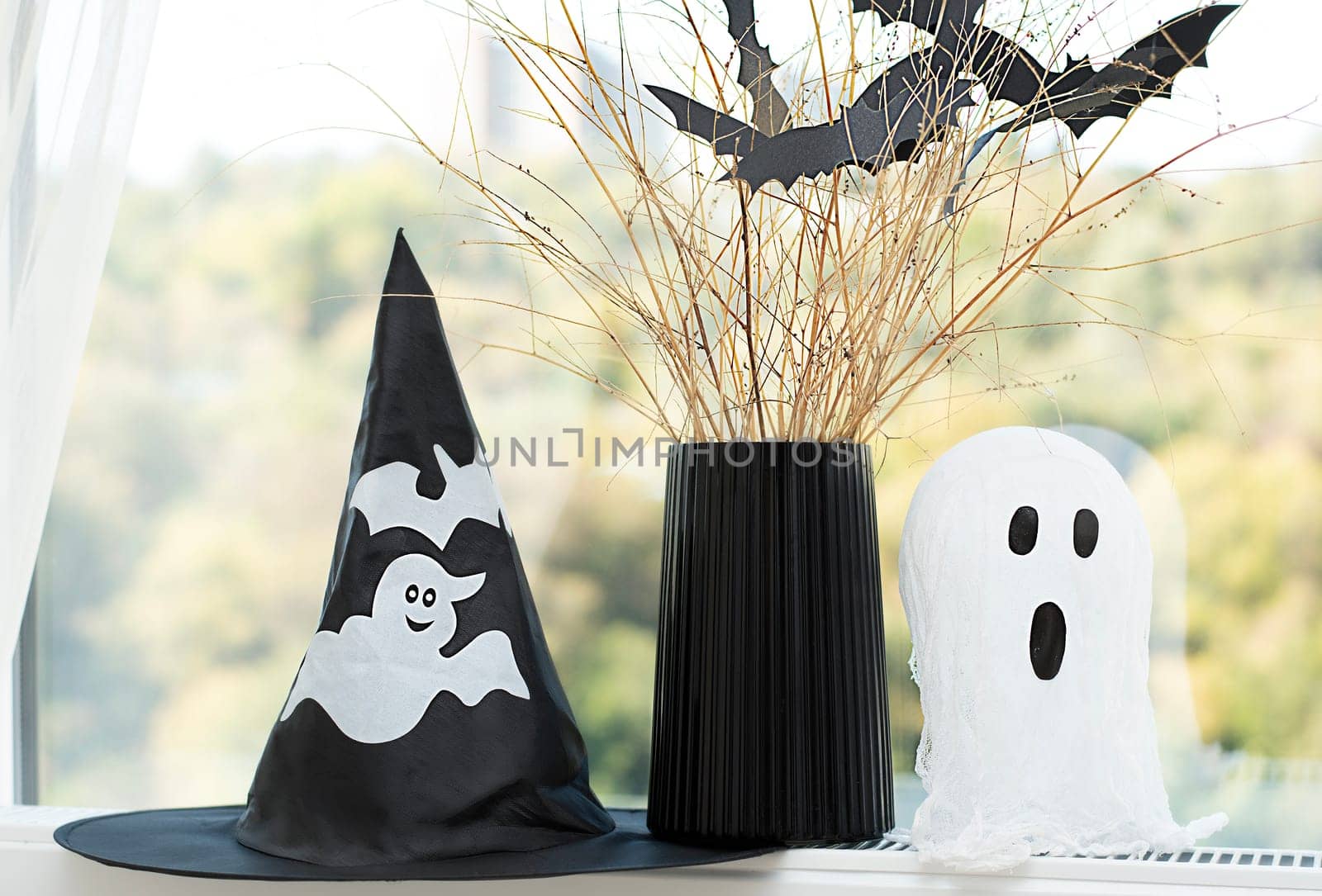 Halloween. Concept. Autumn holiday. A black wizard's hat, a white ghost and a black vase with bats made of paper against the background of a window. Layout. Background, Close-up.