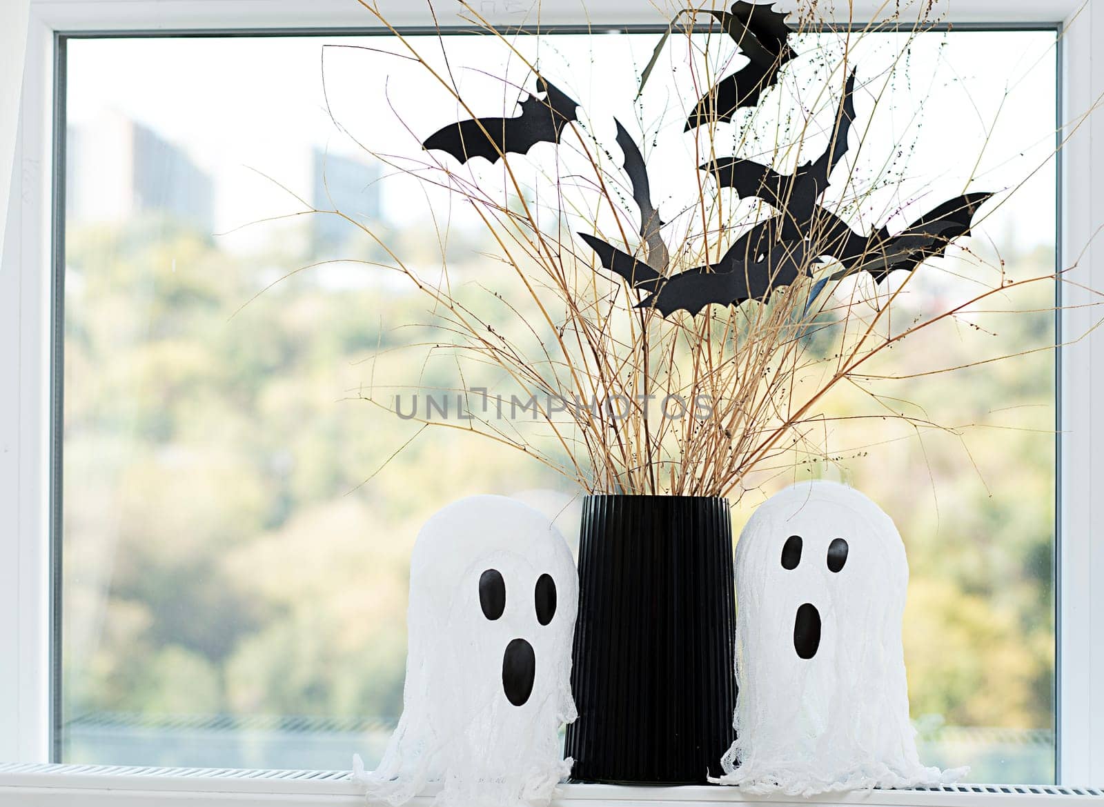 Halloween. Concept. Autumn holiday. Layout. Background.Two white ghosts and a black vase with dry branches and bats made of paper against the background of a window. Close-up.
