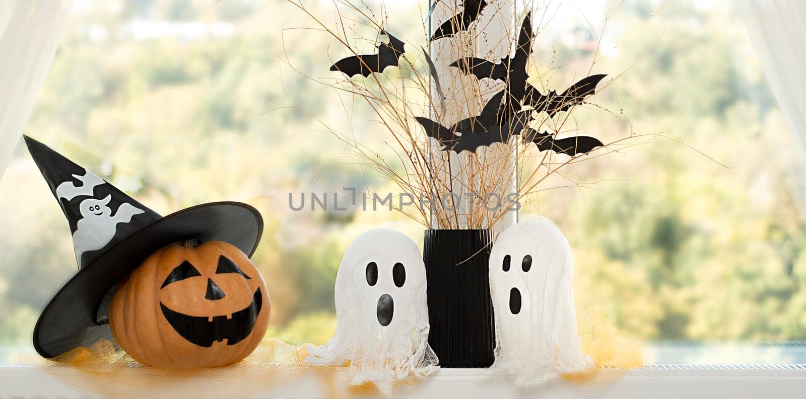 Halloween. Concept. Autumn holiday. Layout. Background. An orange pumpkin in a wizard's hat, two white ghosts and a black vase with dry branches and paper bats on the background of a window. Close-up.