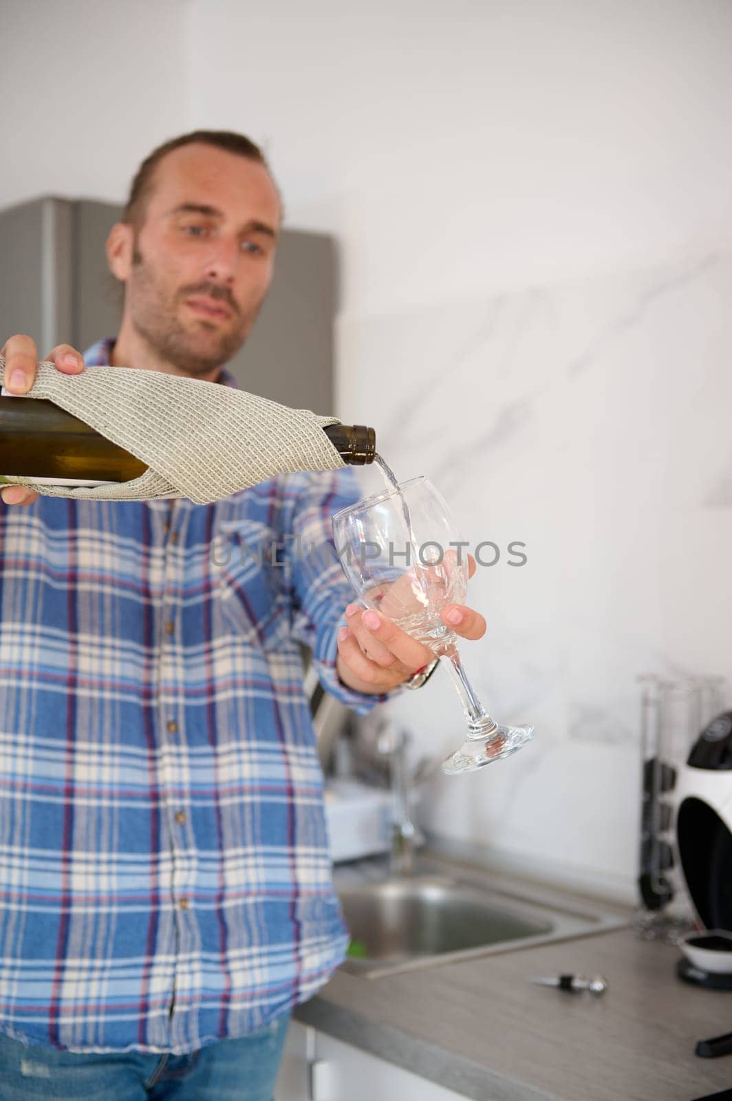 Details on the hands holding bottle of white wine and pouring it into a wine glass, standing in the home kitchen interior by artgf