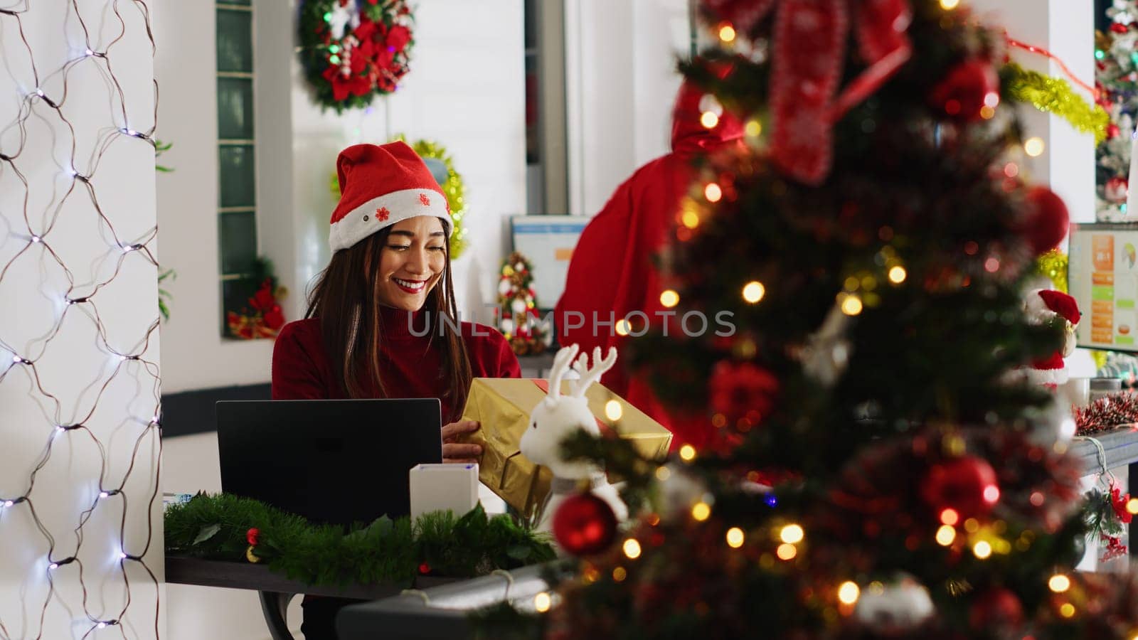 Asian worker receiving gifts from generous colleague acting as Santa in xmas decorated office. Employee wearing festive costume surprising coworkers with presents during winter holiday season