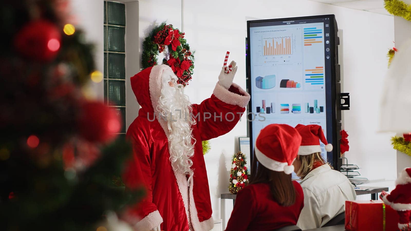 Manager dressed as Santa during Christmas season holding team meeting, showing next year company strategy on digital screen. Team leader in festive decorated office talking with employees