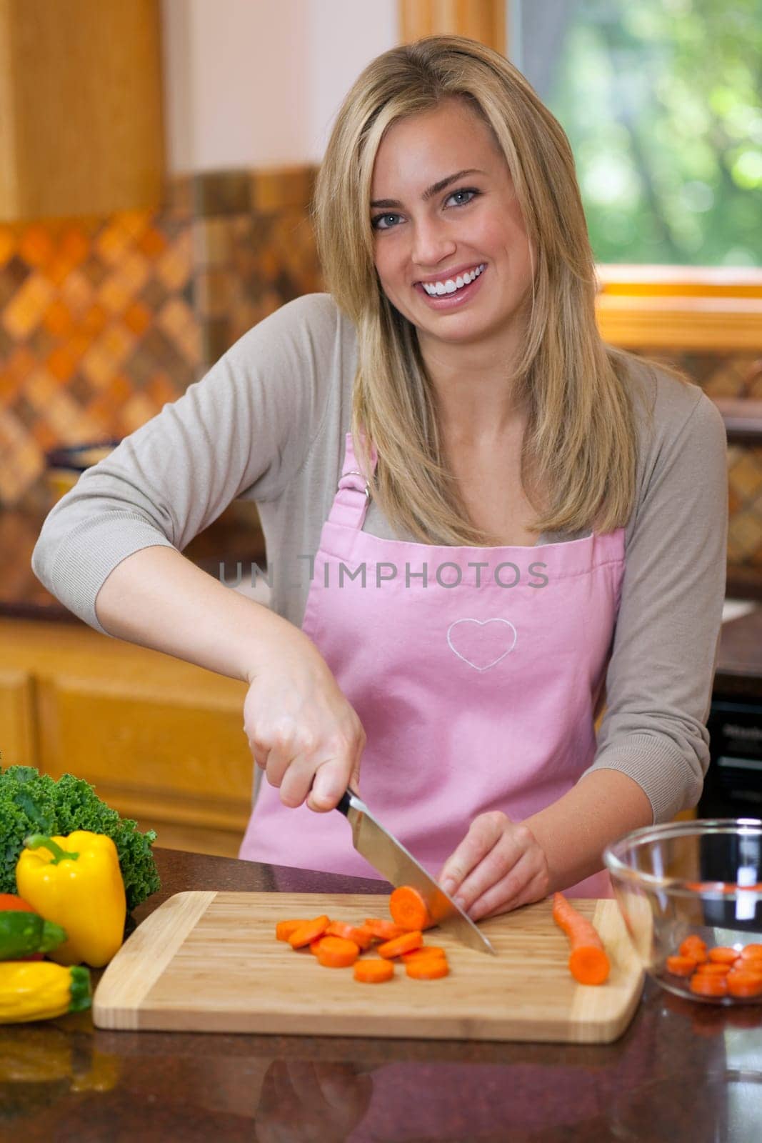 A young woman is slicing carrots in her kitchen and smiling at the camera.  Vertically framed shot.