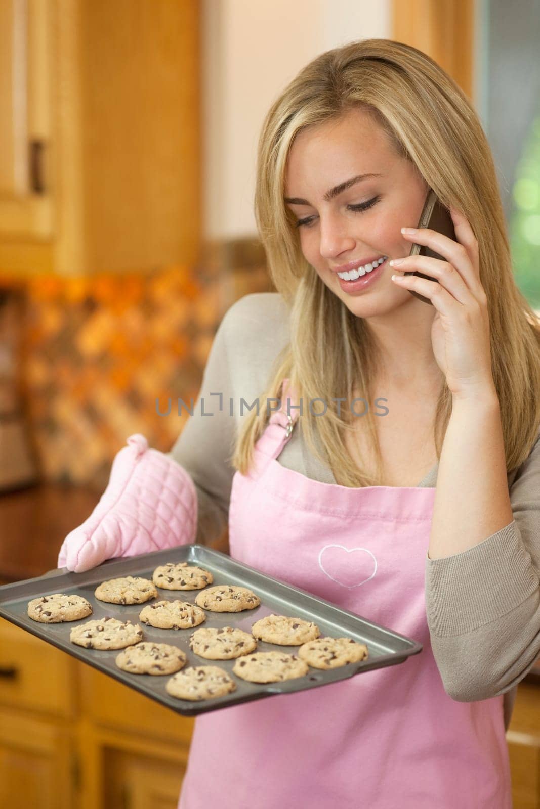 A young woman is holding a baking sheet of cookies, smiling, and talking on the phone.  Vertically framed shot.