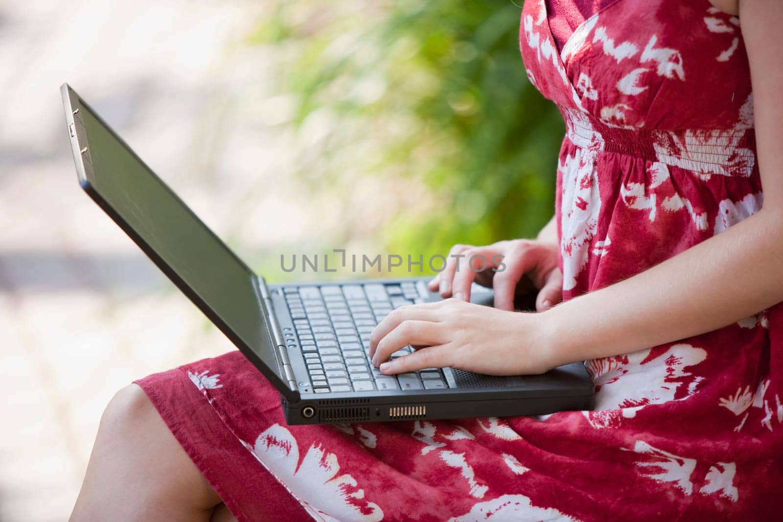 Cropped view of a woman using a laptop in an outdoor setting. She is viewable from neck to knees. Horizontal format.