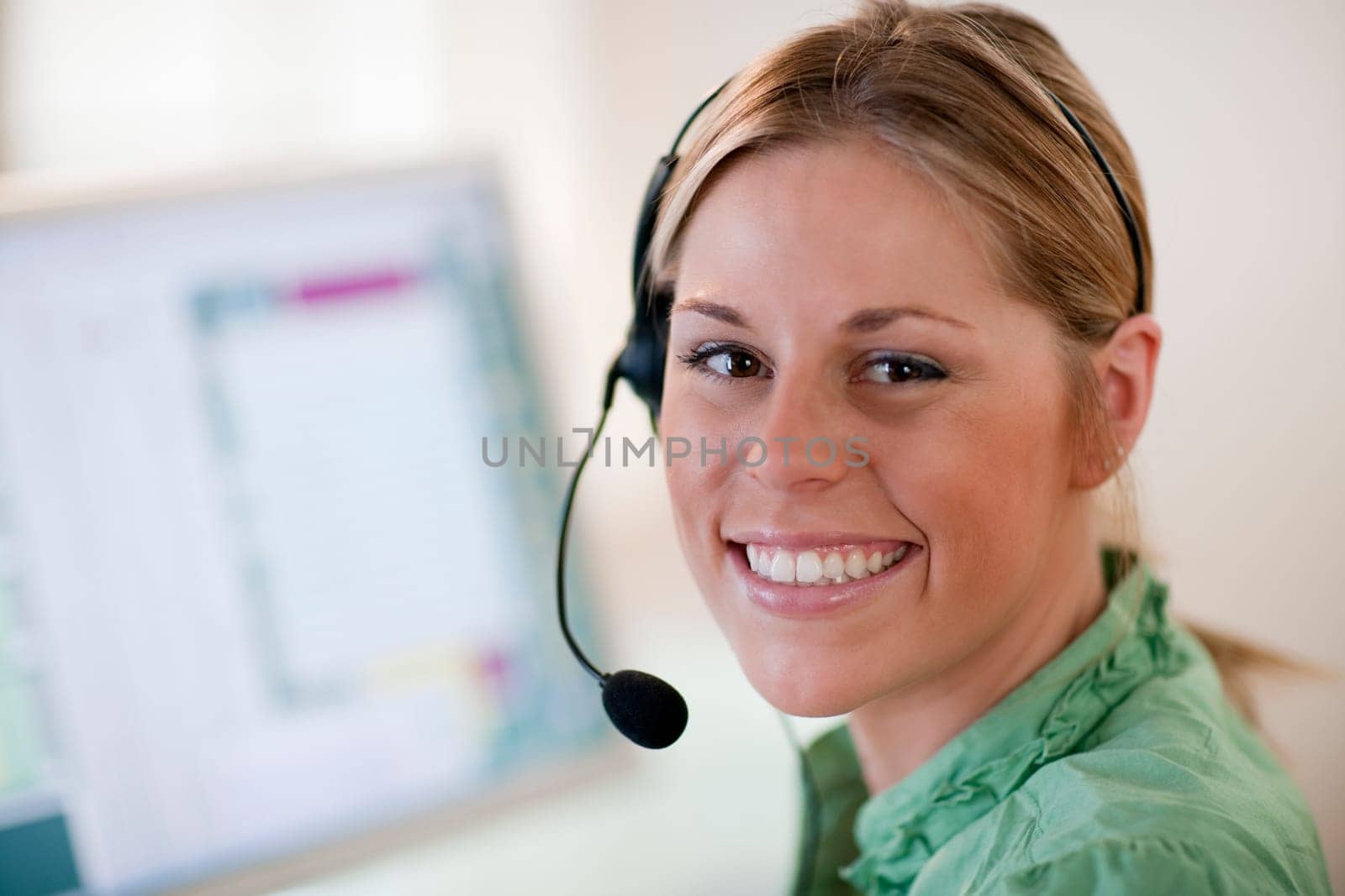 Close-up of a young woman in front of a computer, wearing a headset and smiling at the camera. Horizontal format.