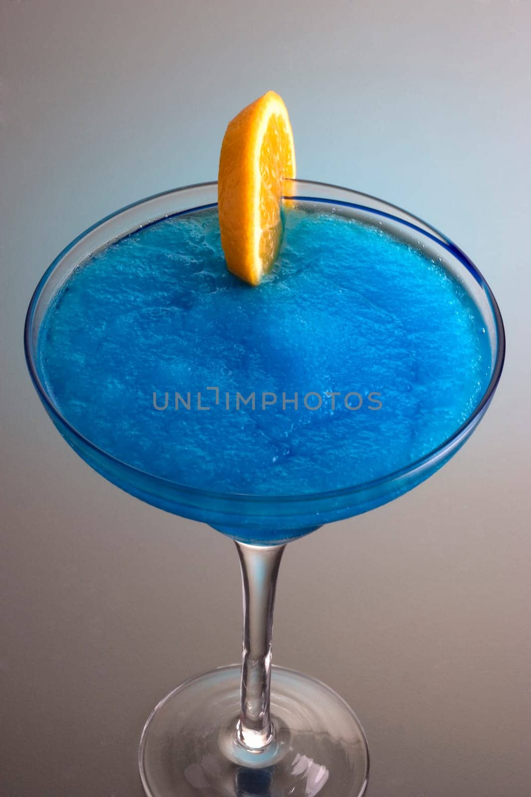 Frozen Blue Hawaiian mixed drink with orange slice garnish on grey background with reflection