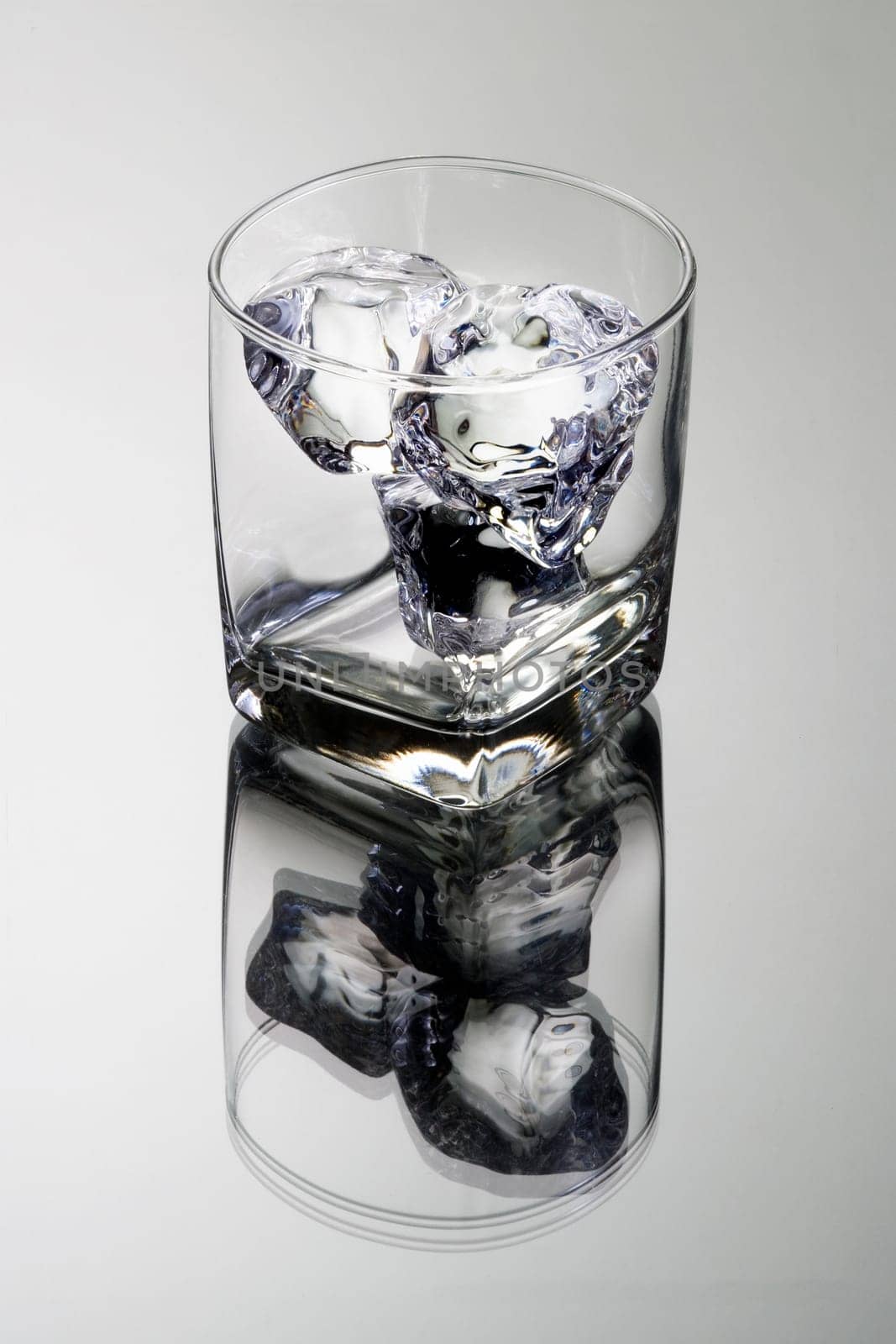 Empty glass tumbler with ice cubes on grey background with reflection