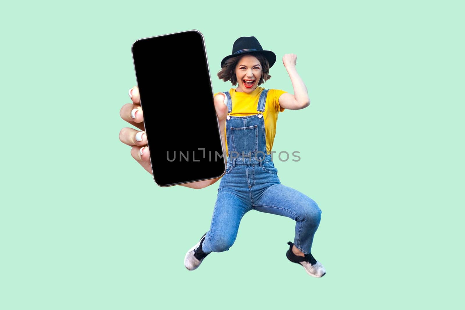 Portrait of happy surprised woman jumping, celebrating her victory, showing big mobile phone with black blank screen, copy space for promotion. Indoor studio shot isolated on light green background.