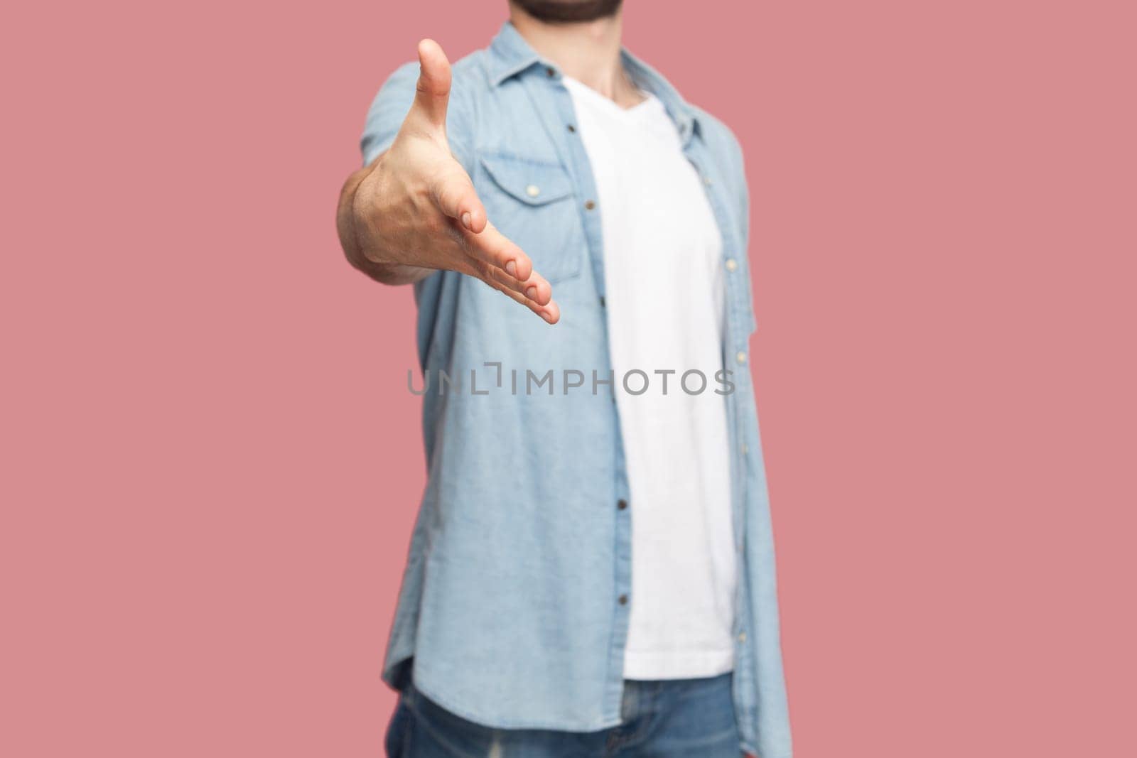 Unknown man in blue casual style shirt standing giving hand for handshake as sign of greeting, saying hello to person he meeting. Indoor studio shot isolated on pink background.