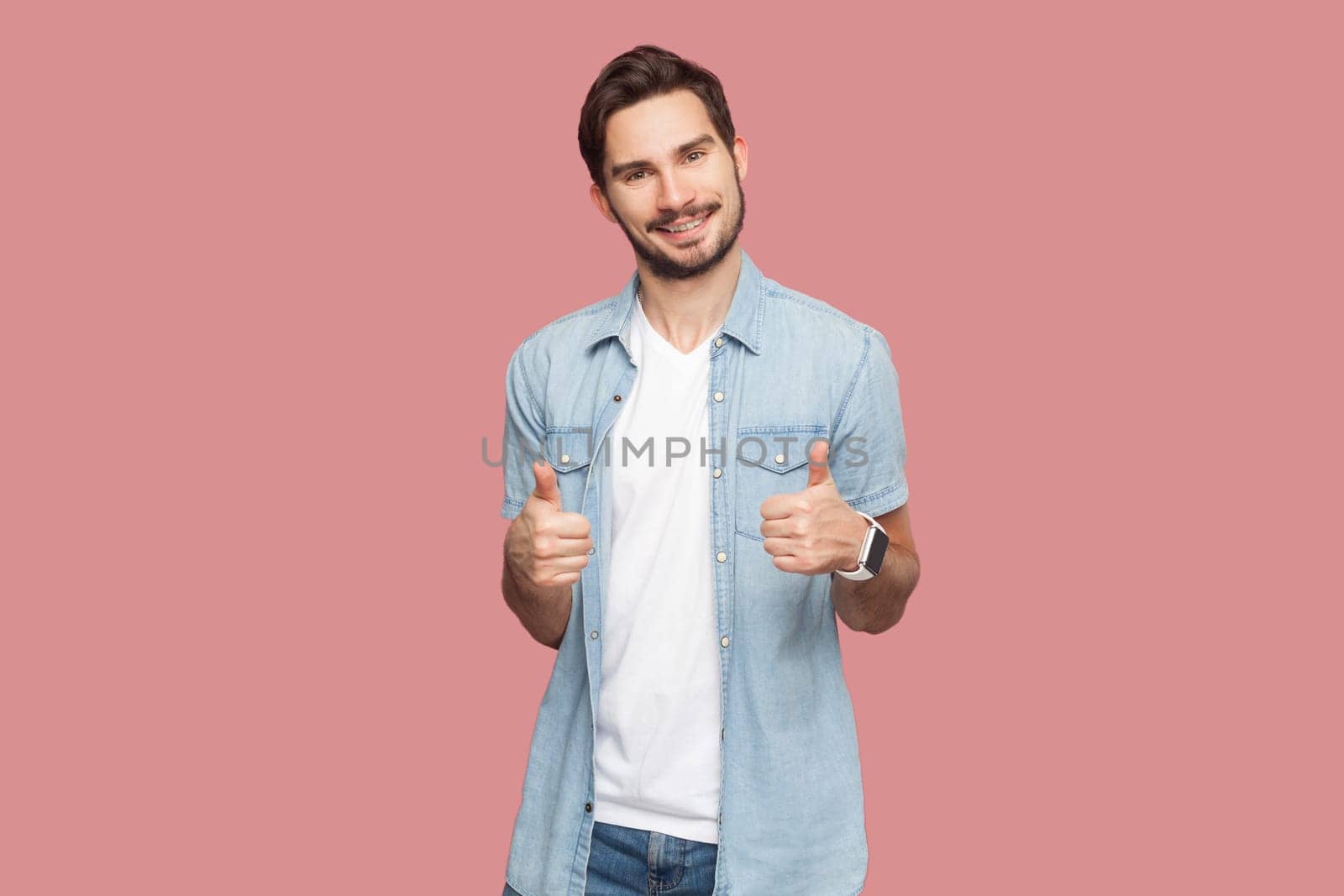 Man in blue shirt gesturing thumbs-up, telling you that you are doing a good job, looking and smiling at the camera with cheerful expression. Indoor studio shot isolated on pink background.