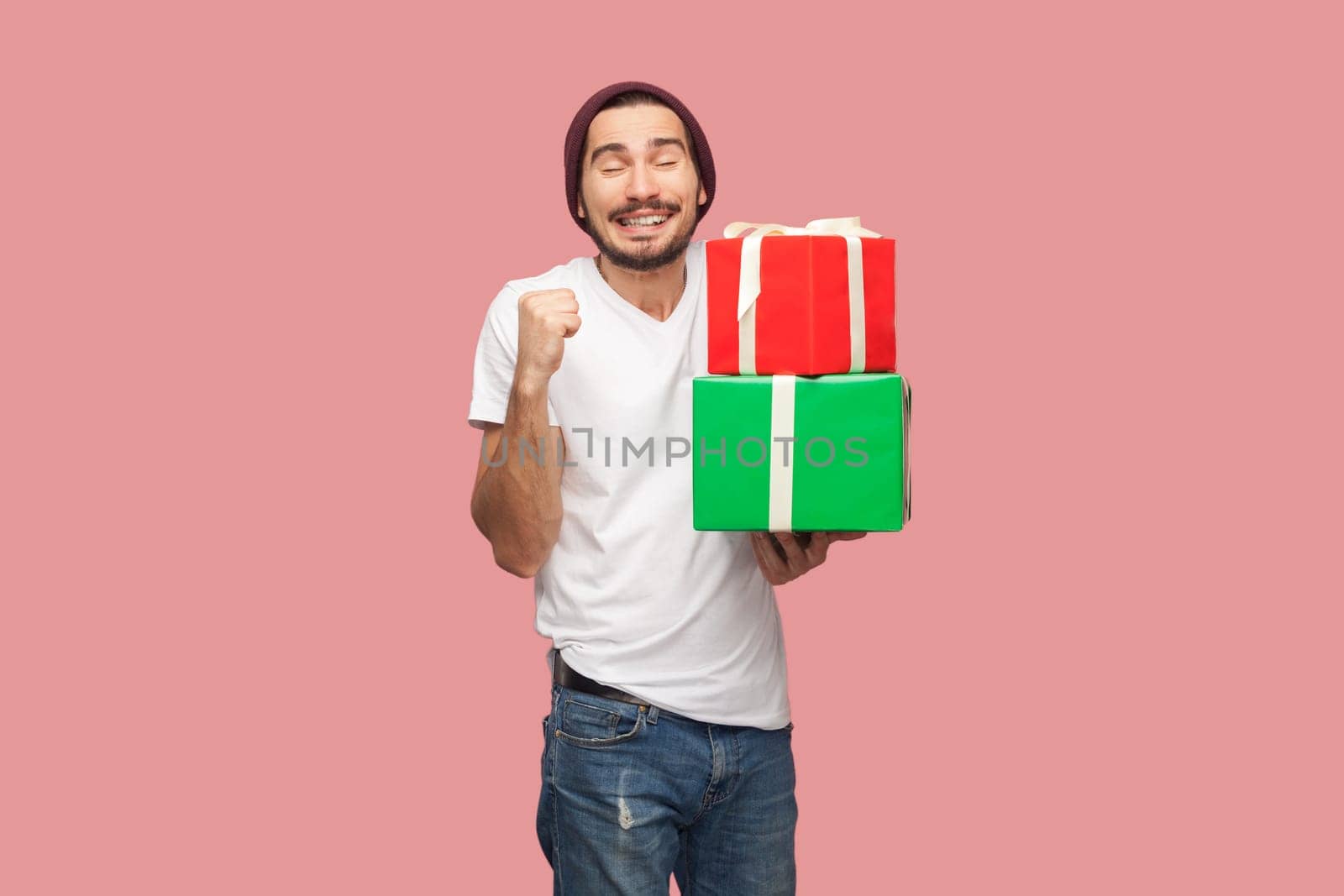 Portrait of satisfied extremely happy bearded man in white T-shirt and beany hat standing holding present box, clenched fist, celebrating. Indoor studio shot isolated on pink background.