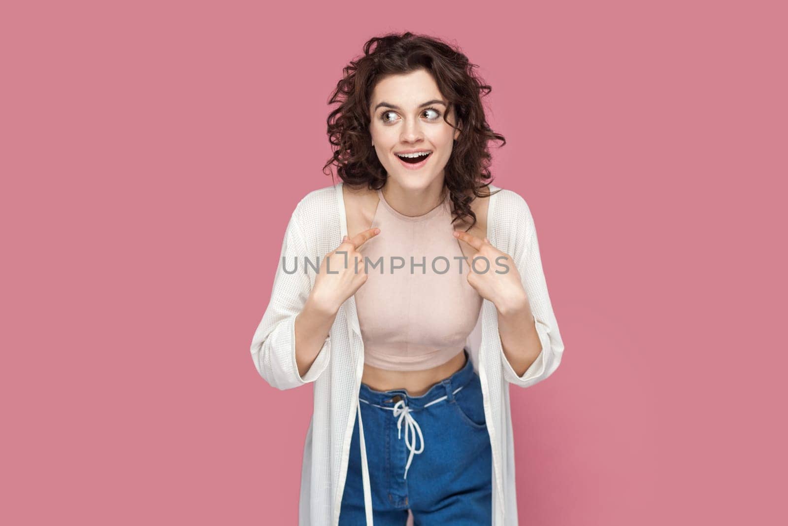 Portrait of excited amazed shocked woman with curly hair wearing casual style outfit pointing both index finger to herself, being surprised to be chosen. Indoor studio shot isolated on pink background