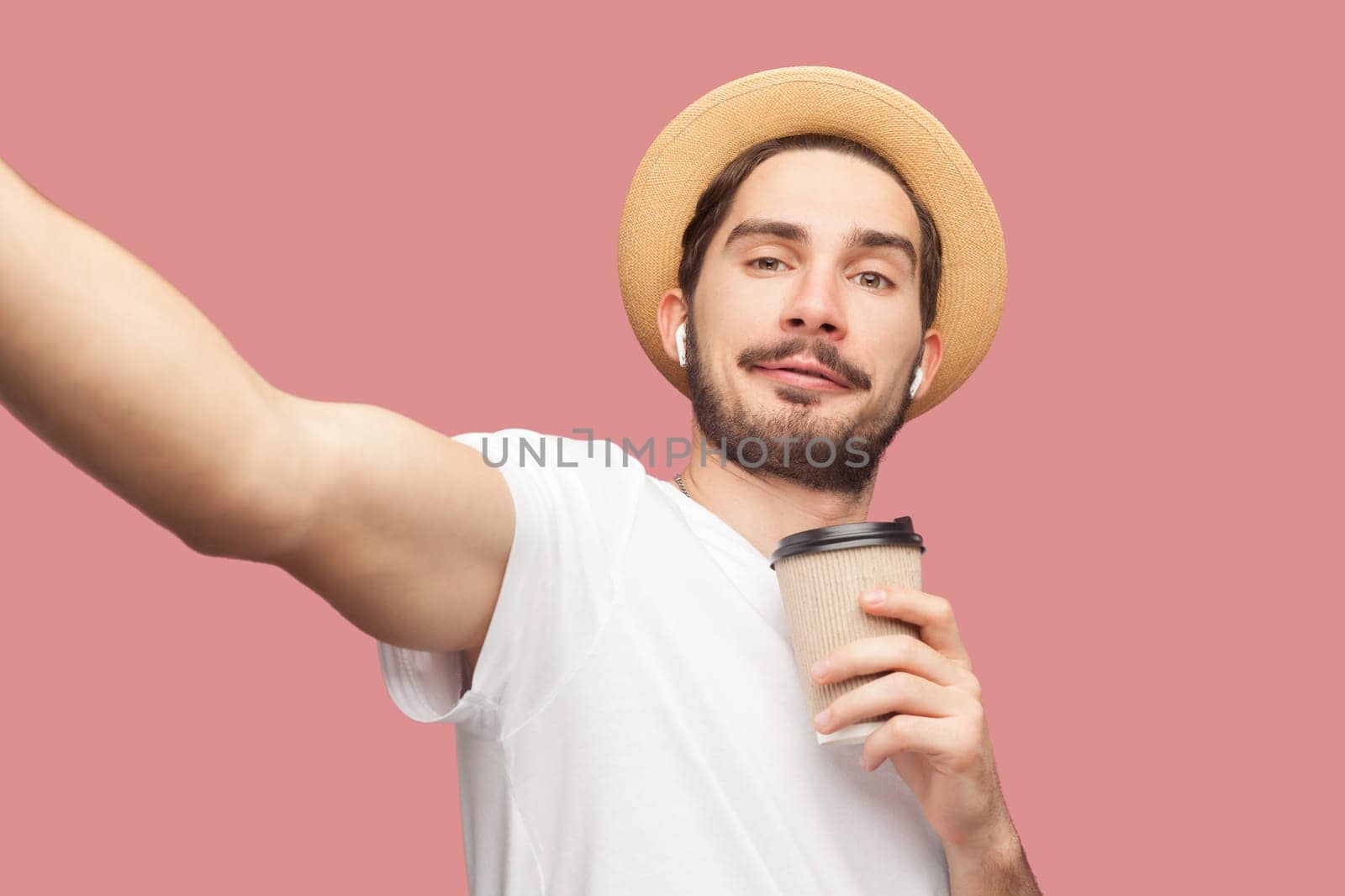 Smiling positive man blogger in T-shirt and hat standing with coffee and making pointy of view photo by Khosro1