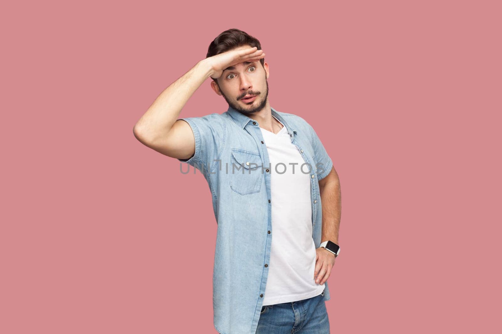 Bearded man in blue casual style shirt standing keeps palm near forehead, looks with concentrated expression into distance, sees something aside. Indoor studio shot isolated on pink background.