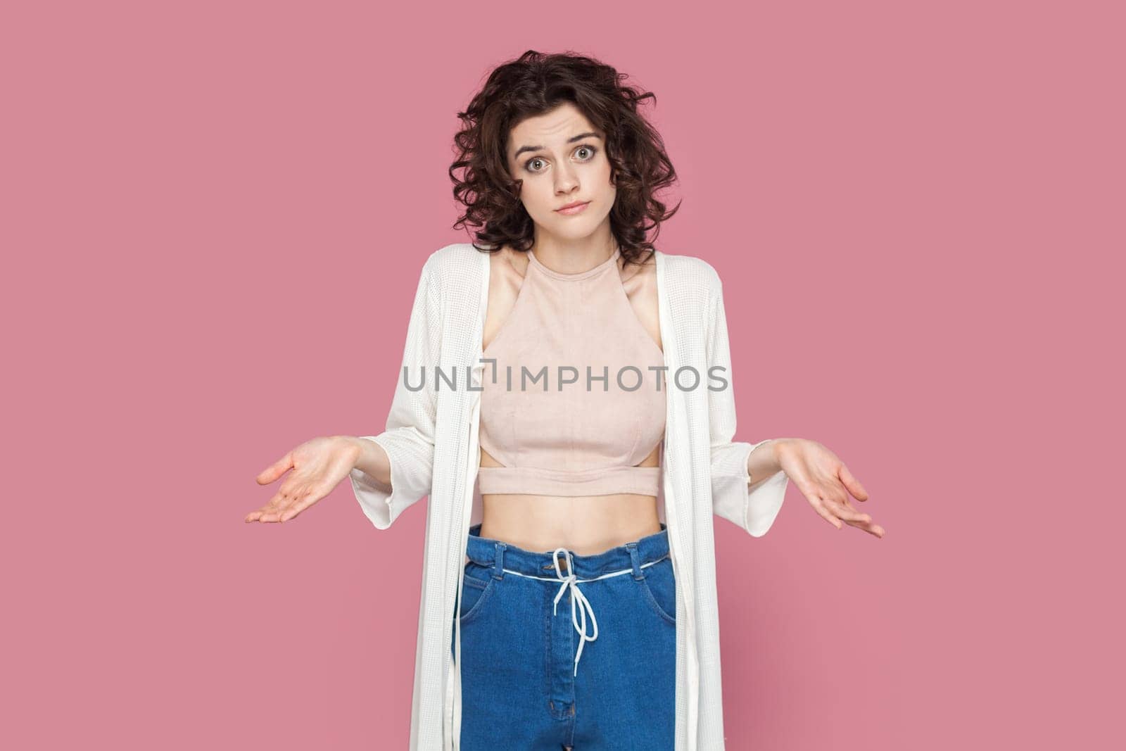 Portrait of puzzled uncertain woman with curly hairstyle wearing casual style outfit spreads hands aside, doesn't know answer. Indoor studio shot isolated on pink background.