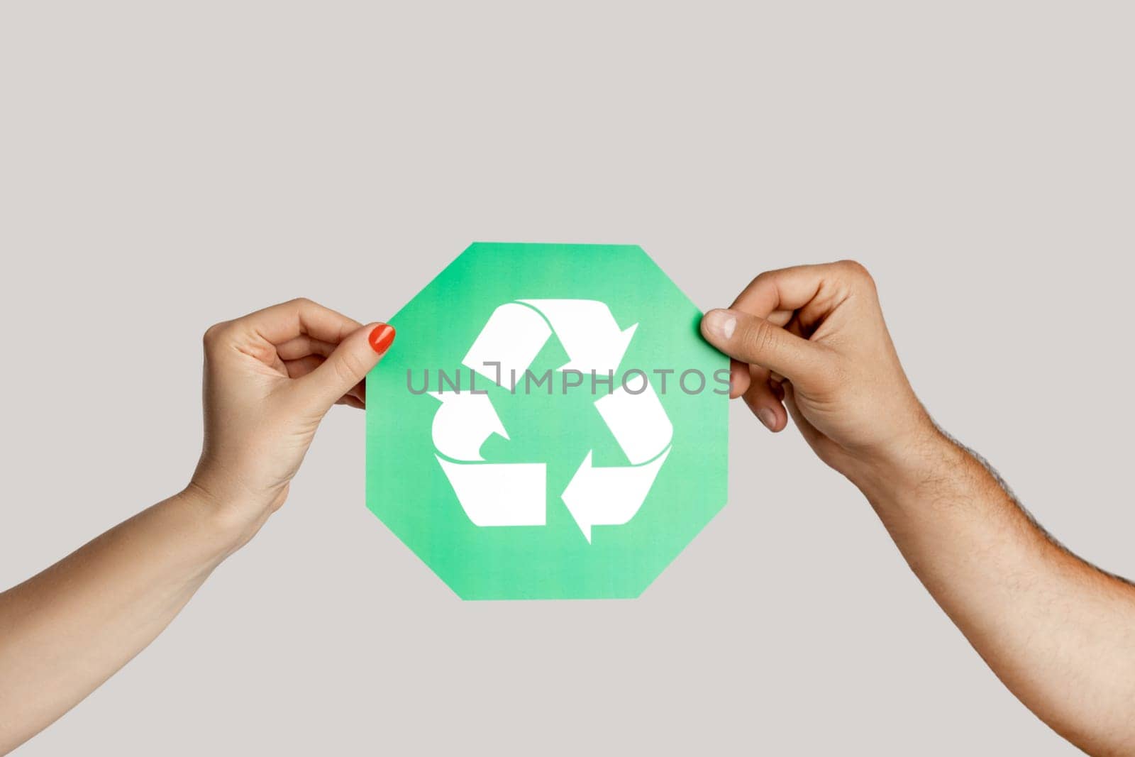 Closeup of woman and man hand holding green recycling sign, ecological icon, environment protection. Indoor studio shot isolated on gray background.