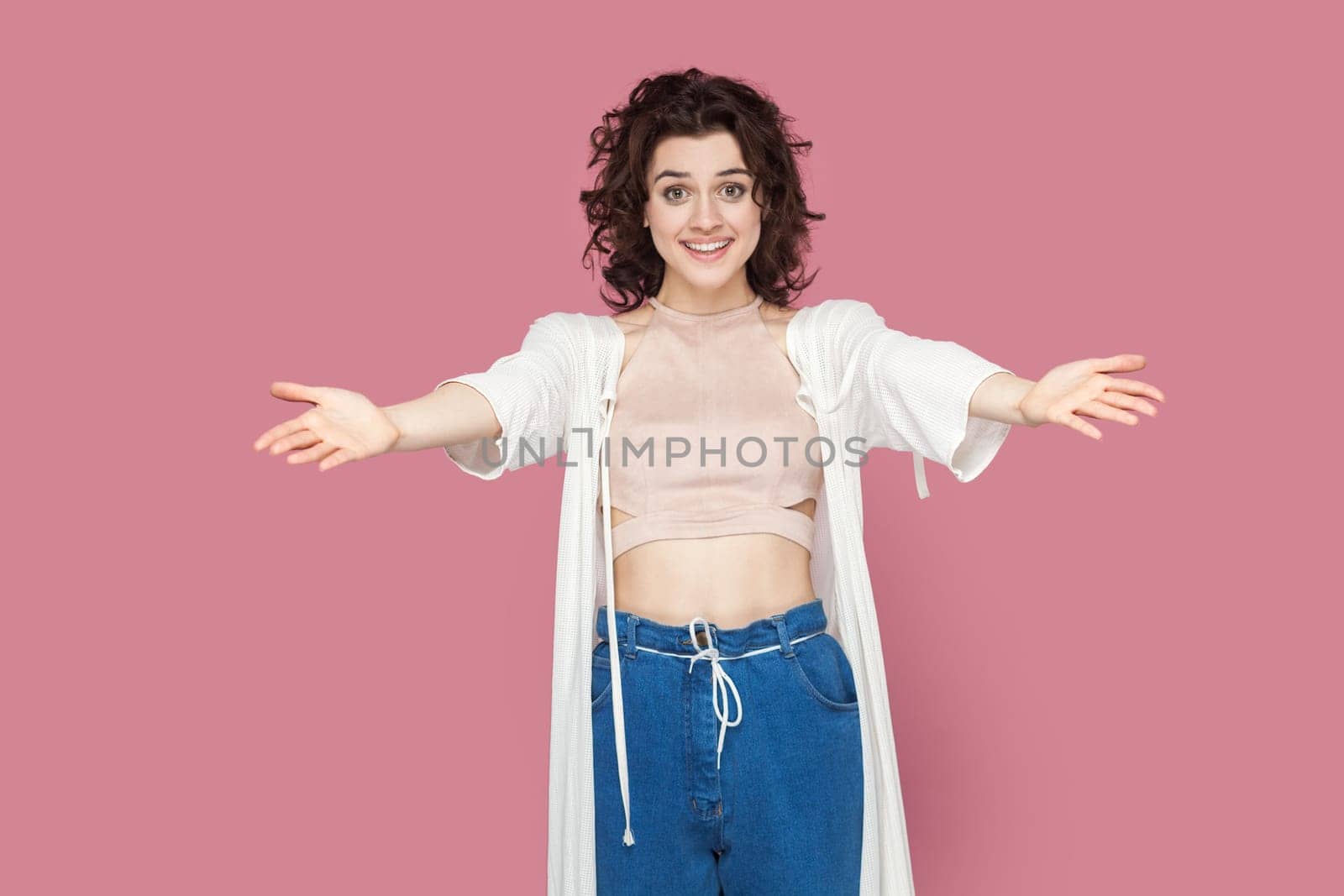 Welcome. Portrait of extremely happy pretty woman with curly hair wearing casual style outfit standing with outstretched hands, inviting. Indoor studio shot isolated on pink background.