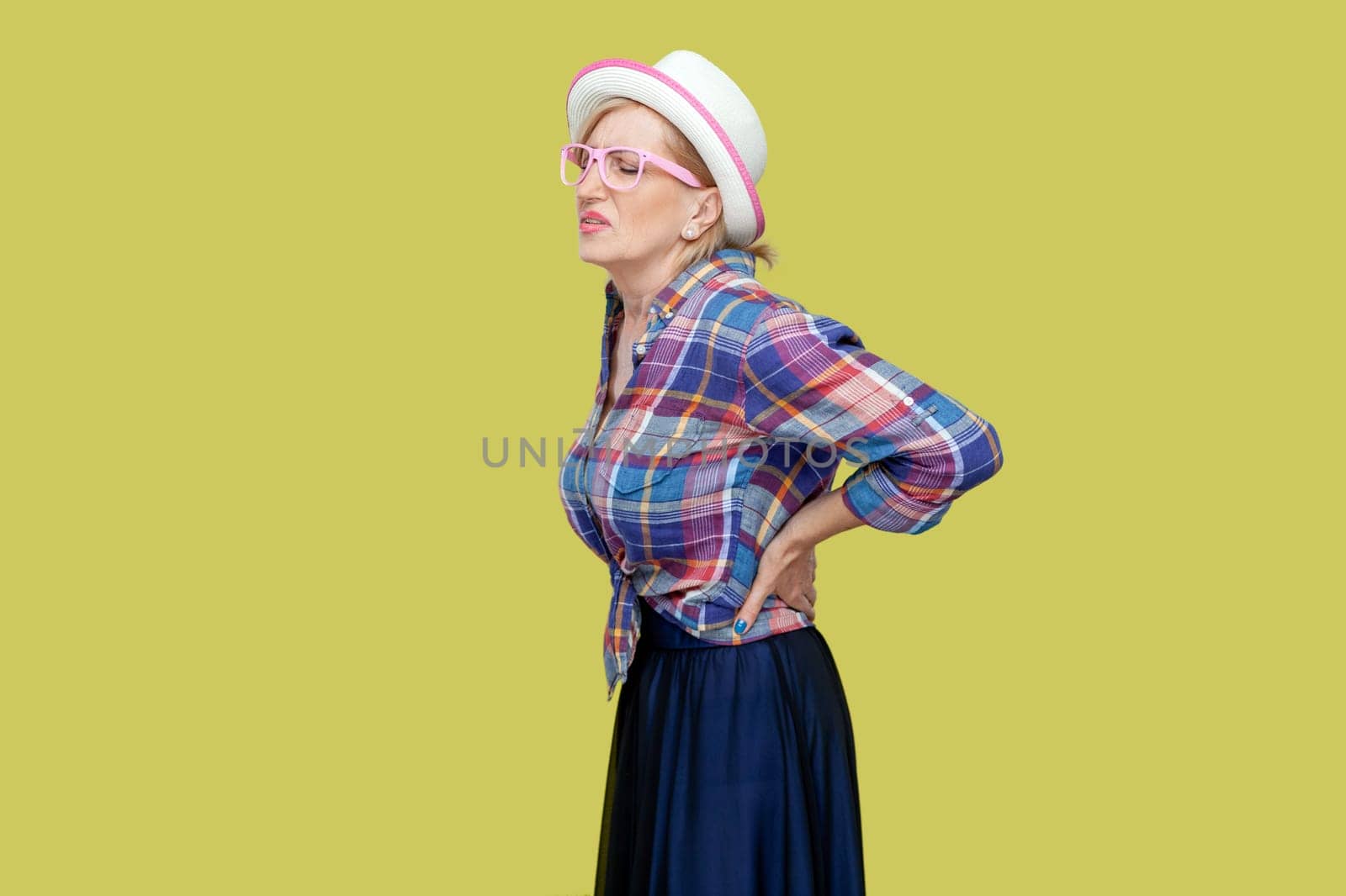 Senior woman wearing checkered shirt, hat and eyeglasses suffers from radiculitis, keeps hand on waist, problems with health, has pain in spine. Indoor studio shot isolated on yellow background.