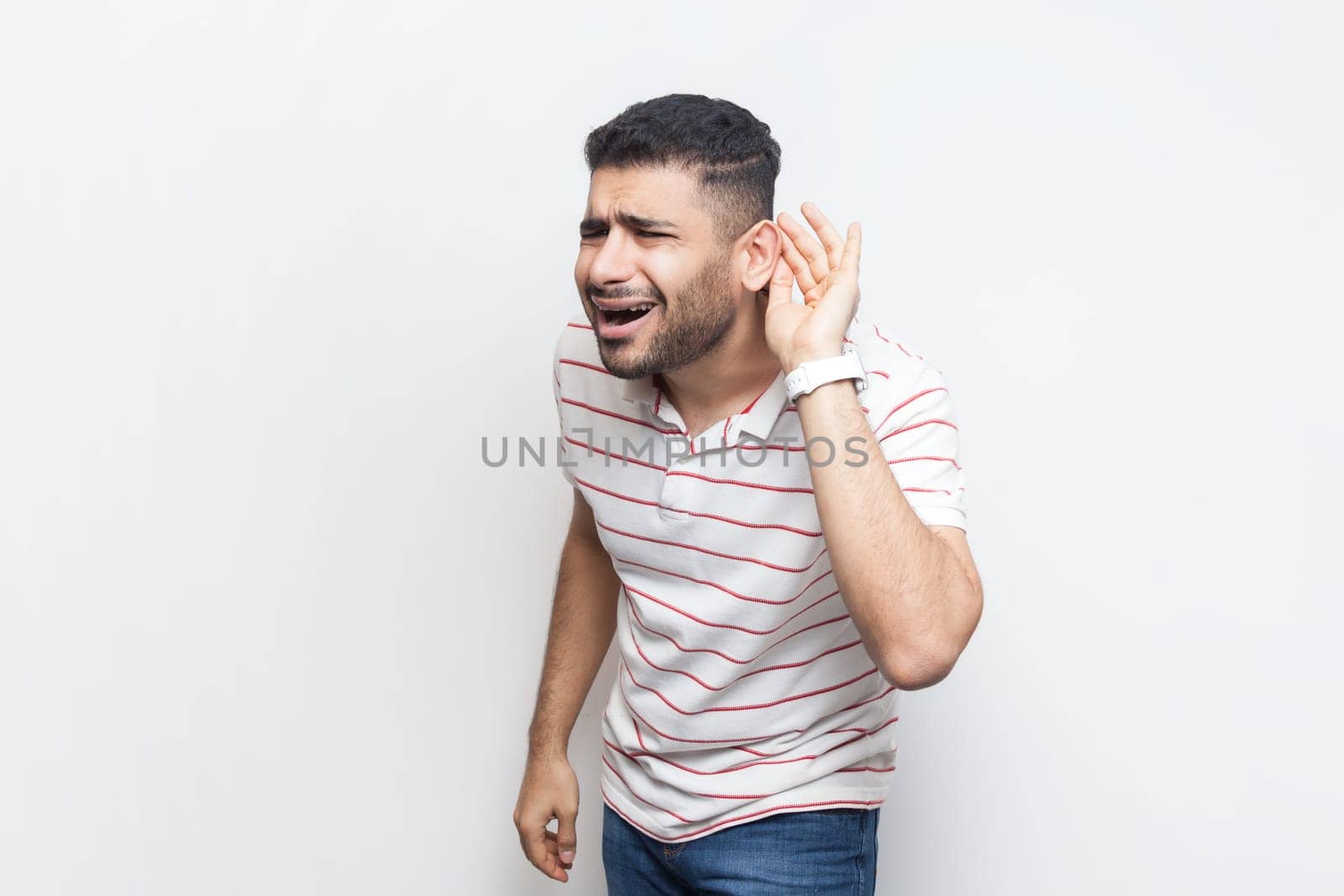 Portrait of attractive handsome bearded man wearing striped t-shirt standing with hands near ear, frowning face, listening silent conversation. Indoor studio shot isolated on gray background.