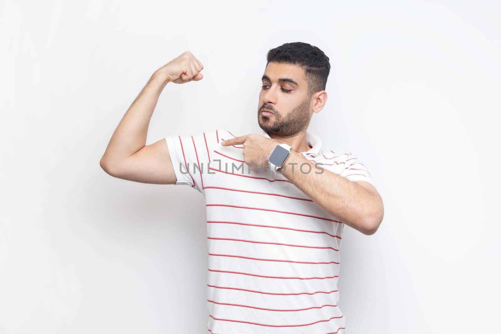 Portrait of strong attractive bearded man wearing striped t-shirt standing with raised hand, pointing at his biceps, looks proud. Indoor studio shot isolated on gray background.