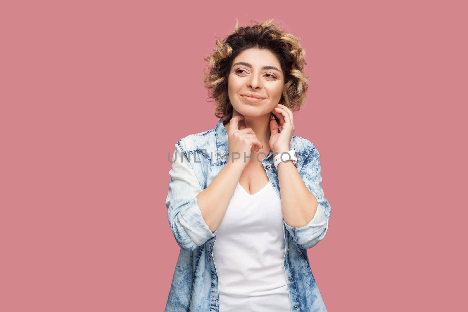 Portrait of smiling beautiful woman with curly hairstyle wearing blue shirt standing looking away, smiling, dreaming about something pleasant. Indoor studio shot isolated on pink background.