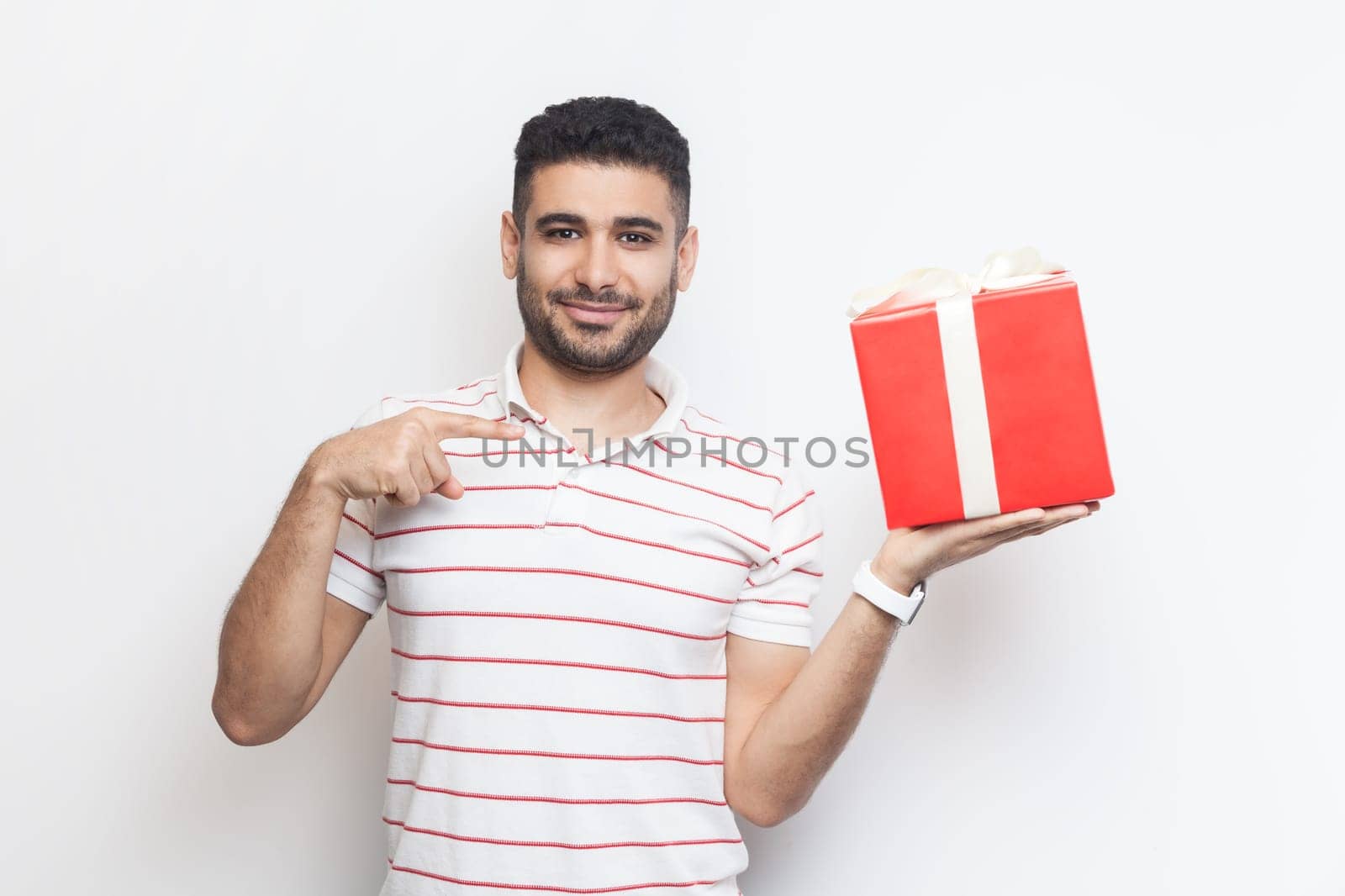 Smiling positive man wearing t-shirt standing holding red wrapped present box, pointing at gift. by Khosro1