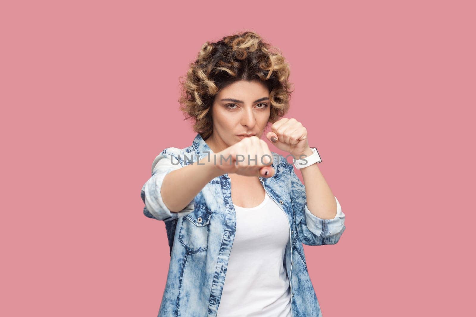 Portrait of angry aggressive woman with curly hairstyle wearing blue shirt standing with clenched fist, boxing, knocks somebody. Indoor studio shot isolated on pink background.