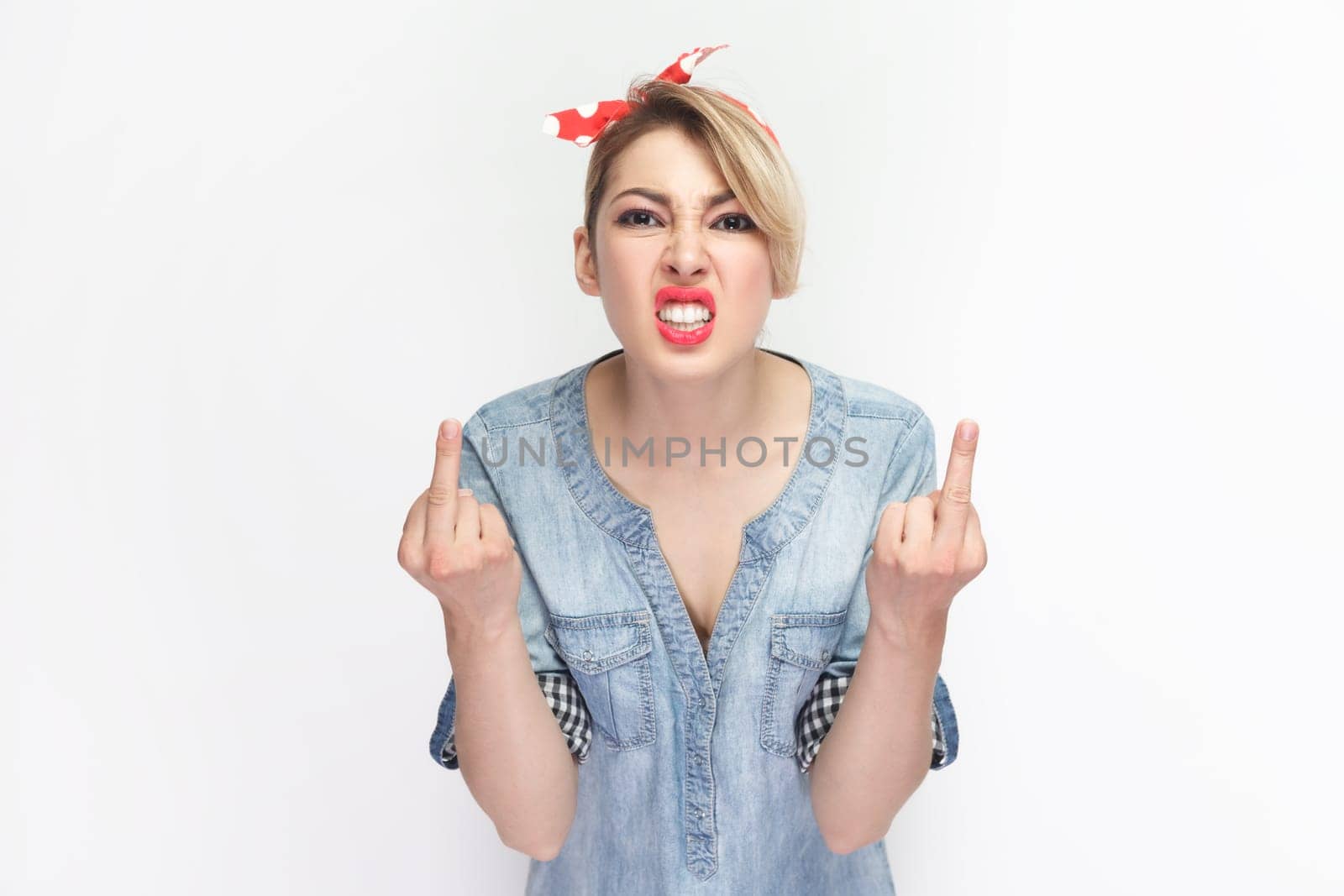 Portrait of angry aggressive rude blonde woman wearing blue denim shirt and red headband standing showing fuck sign, arguing with somebody. Indoor studio shot isolated on gray background.