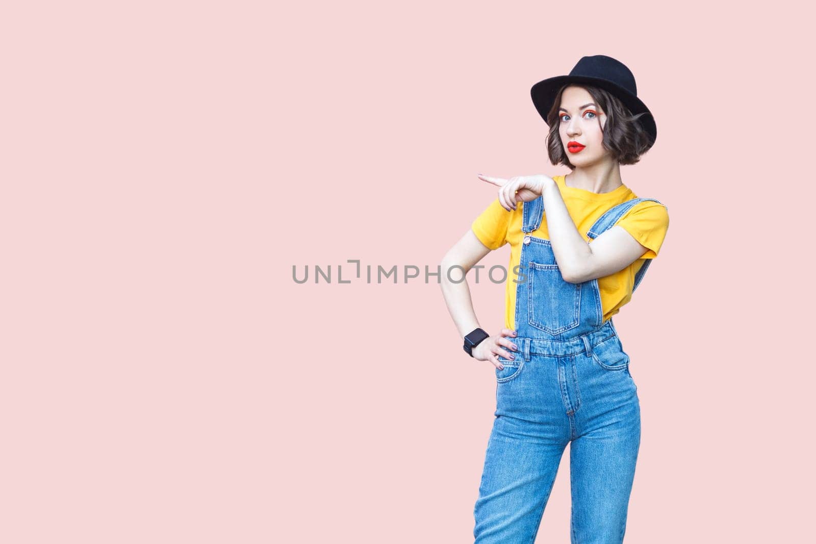 Portrait of surprised amazed hipster woman in blue denim overalls, yellow T-shirt and black hat, pointing at copy space for advertisement. Indoor studio shot isolated on light pink background.