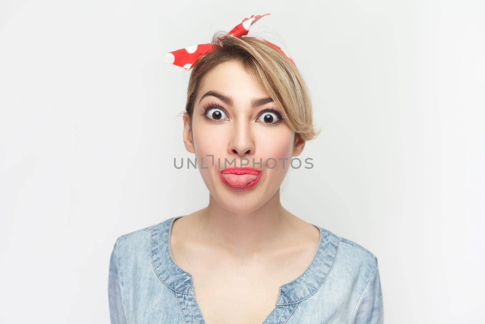 Funny foolish woman in denim shirt and red headband standing sticking tongue out, looking at camera. by Khosro1