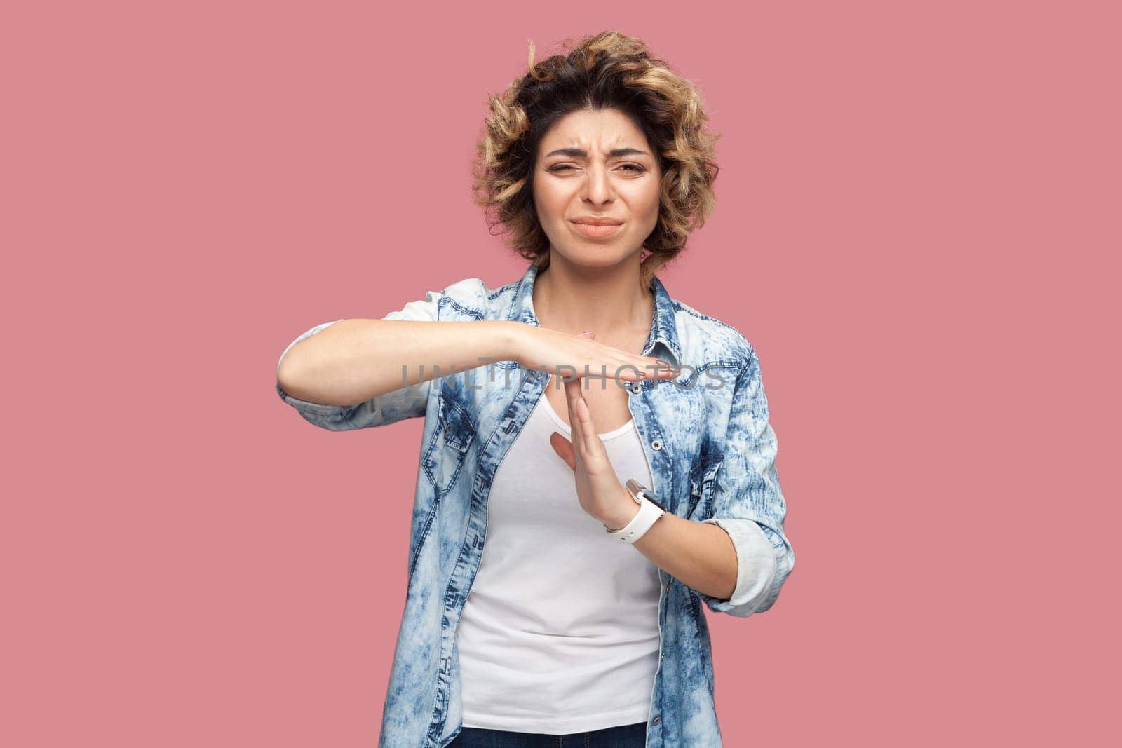 Portrait of sad depressed woman with curly hairstyle wearing blue shirt standing showing time out gesture, looking at camera, expressing sadness. Indoor studio shot isolated on pink background.