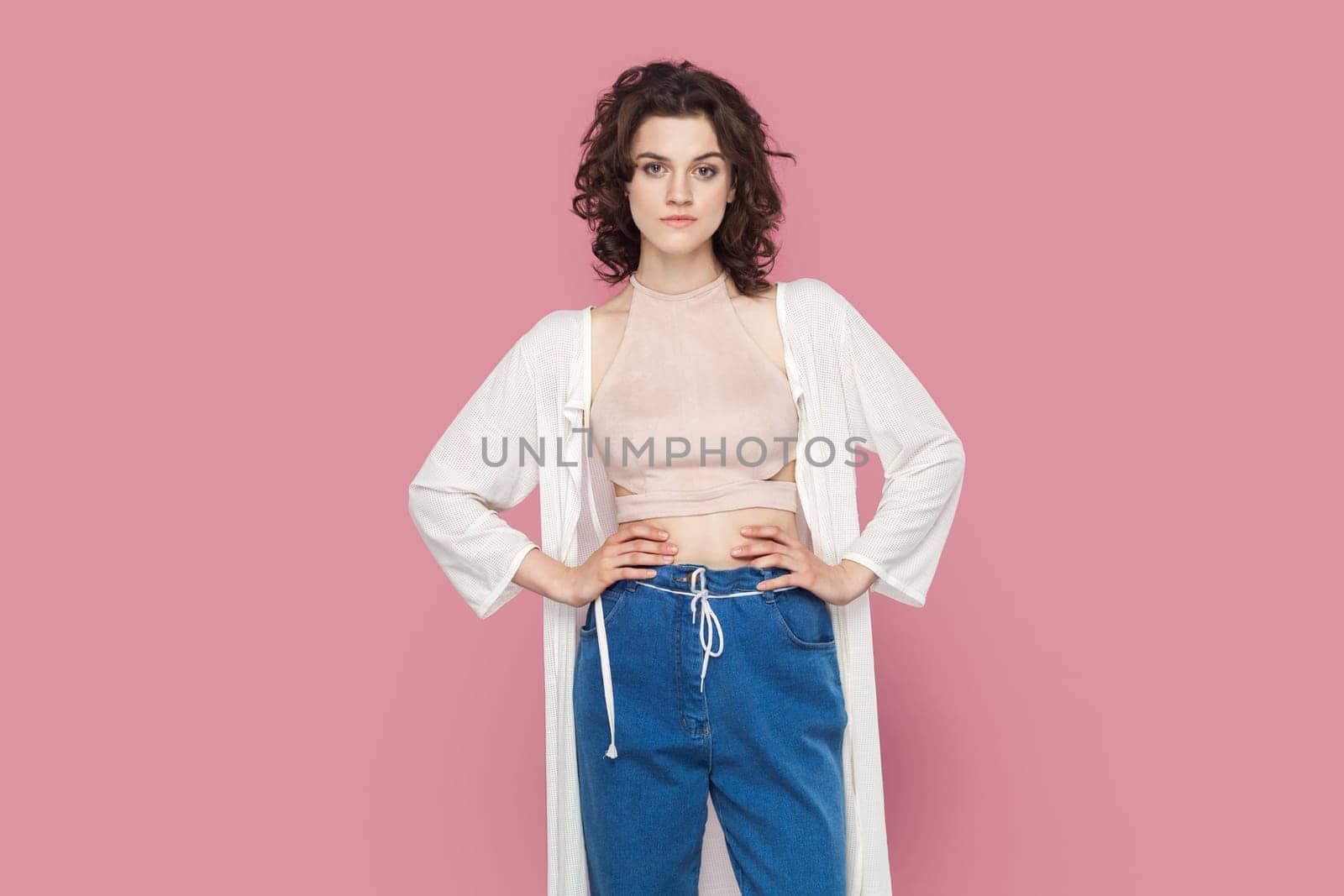 Portrait of self-confident egoistic woman with curly hair wearing casual style outfit standing with hands on hips, looking at camera, feels pride. Indoor studio shot isolated on pink background.