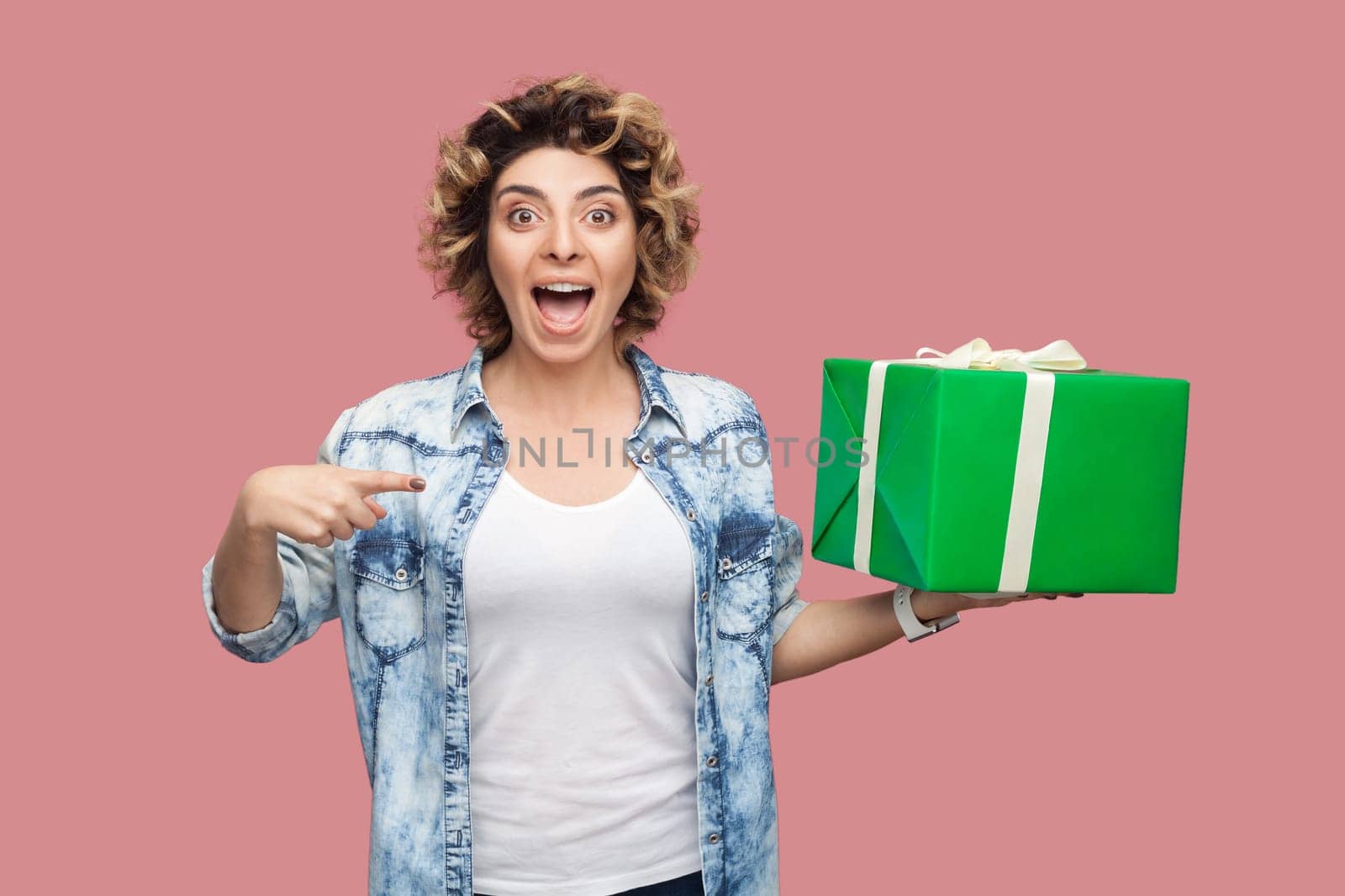 Portrait of excited happy beautiful woman with curly hairstyle wearing blue shirt pointing at green present box, being surprised, celebrating birthday. Indoor studio shot isolated on pink background.