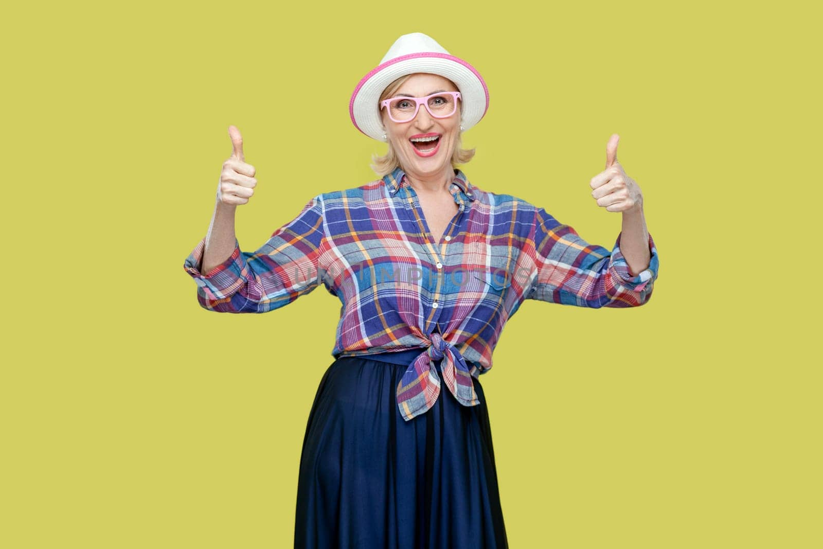 Portrait of senior woman makes thumbs up sign, demonstrates support and respect to someone, smiles pleasantly at camera, achieves desirable goal. Indoor studio shot isolated on yellow background.