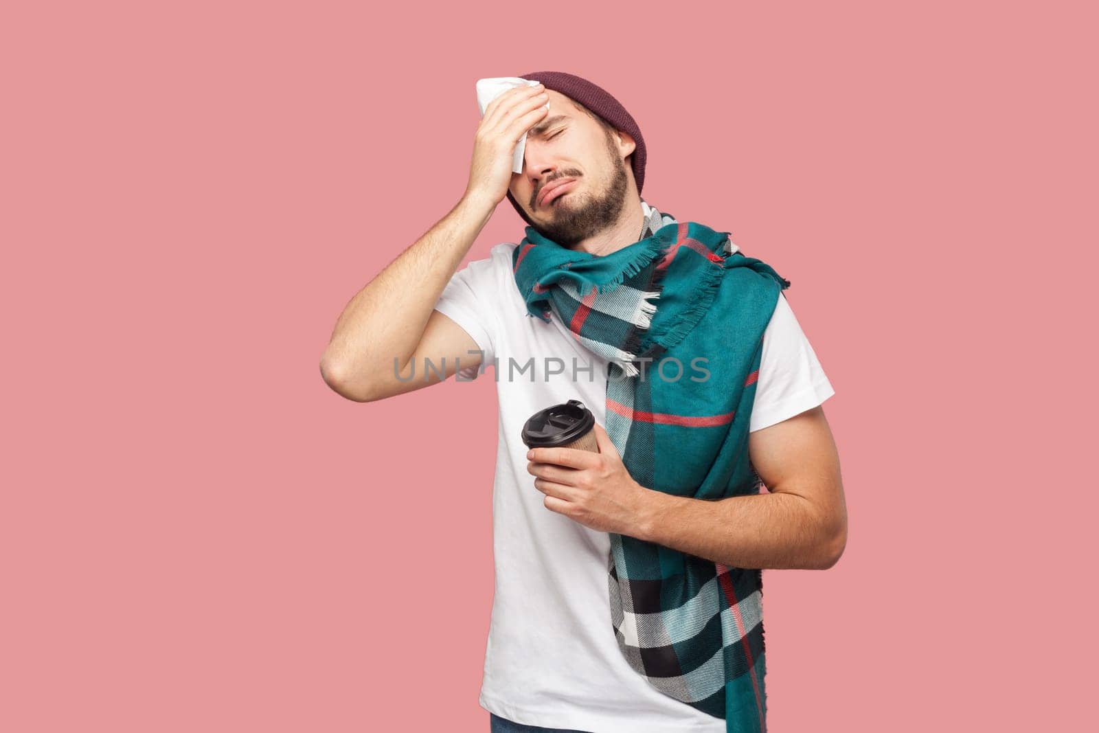 Portrait of sick unhealthy bearded man in white T-shirt and beany hat standing with paper cup and napkin, suffering headache, has high temperature. Indoor studio shot isolated on pink background.