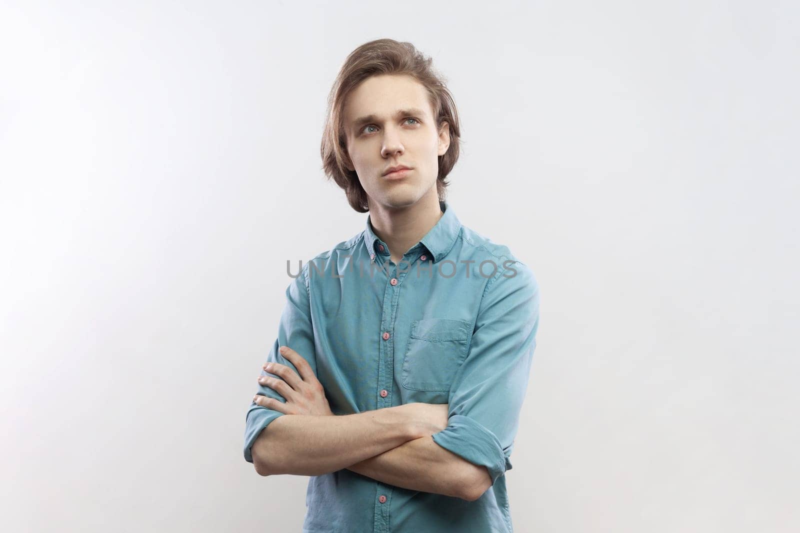 Portrait of pensive thoughtful handsome young man standing with folded hands, thinking, planning, pondering, wearing blue shirt. Indoor studio shot isolated on gray background.