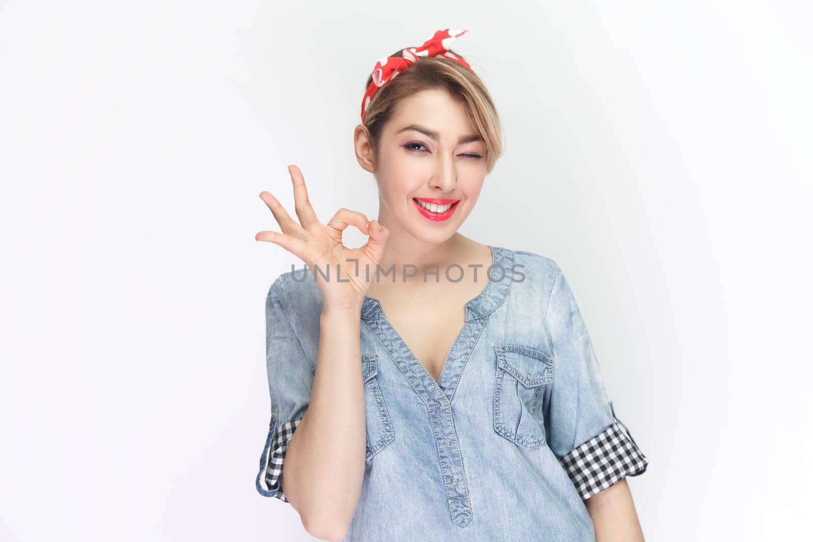 Portrait of blonde woman wearing blue denim shirt and red headband standing making okay gesture, enjoying life, saying ok, confirms information. Indoor studio shot isolated on gray background.