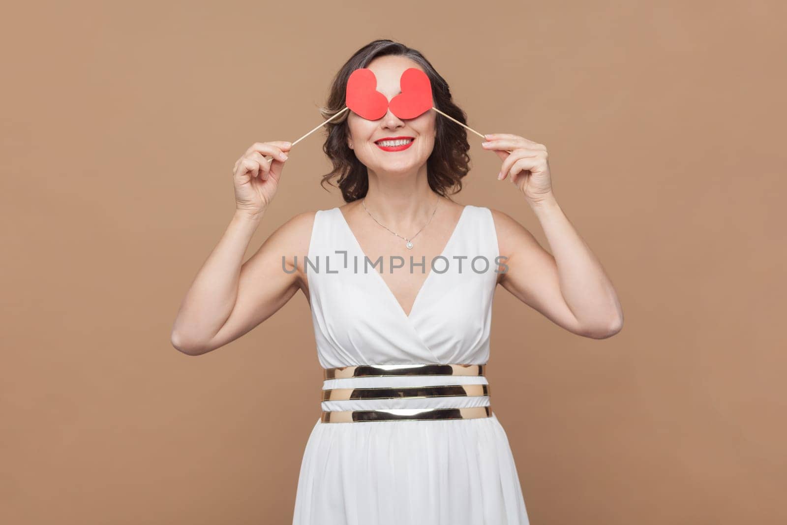 Portrait of romantic smiling woman with wavy hair covering eyes with little red hears, flirting with her boyfriend, wearing white dress. Indoor studio shot isolated on light brown background.