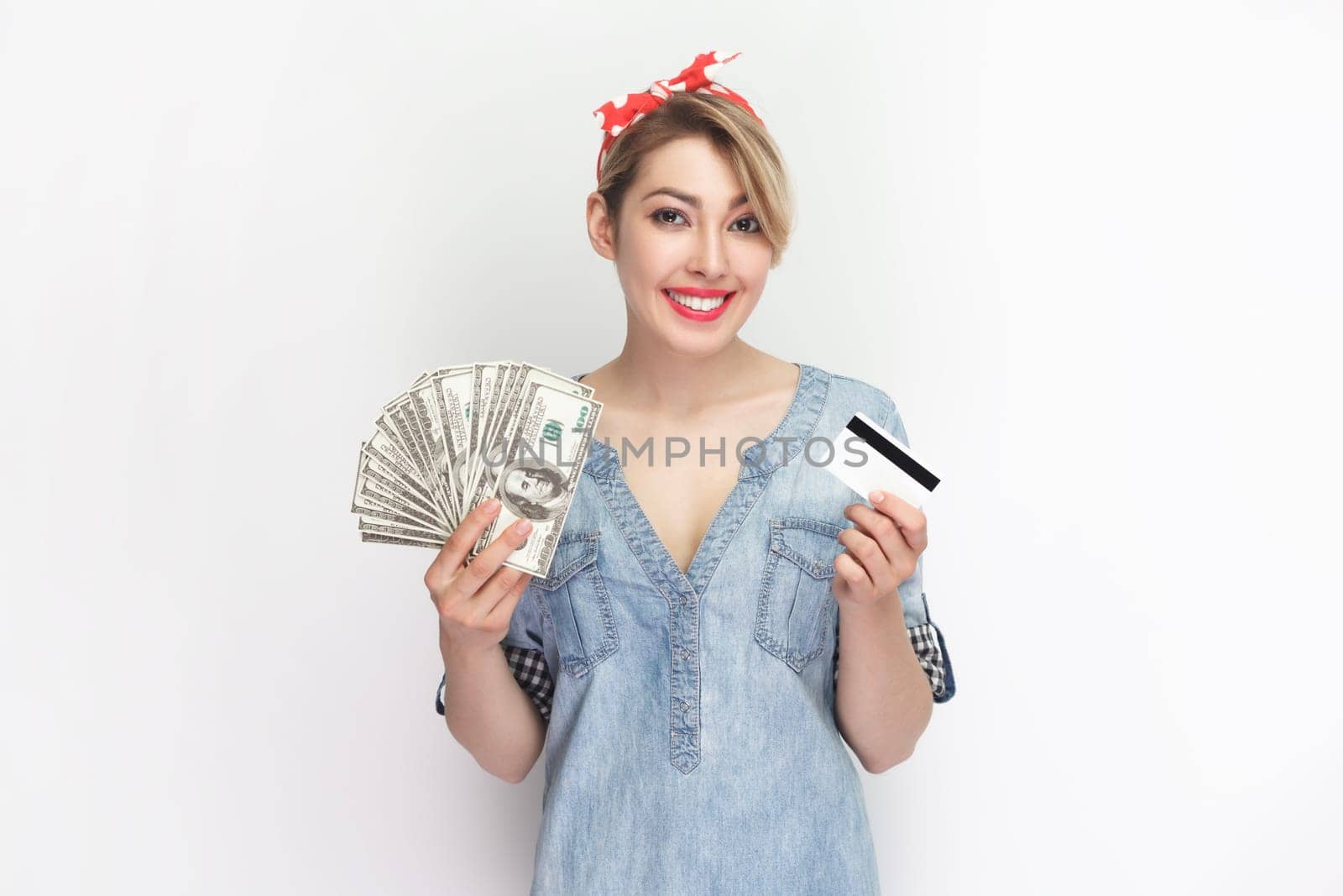 Portrait of smiling attractive cheerful blonde woman wearing blue denim shirt and red headband standing with credit card and dollar banknotes. Indoor studio shot isolated on gray background.