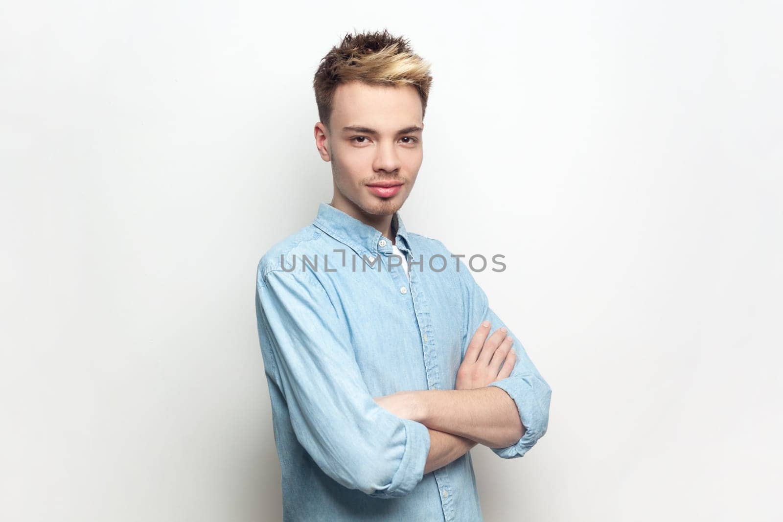 Portrait of self-confident attractive handsome man wearing denim shirt standing with crossed arms, looking at camera, expressing positive emotions. Indoor studio shot isolated on gray background.