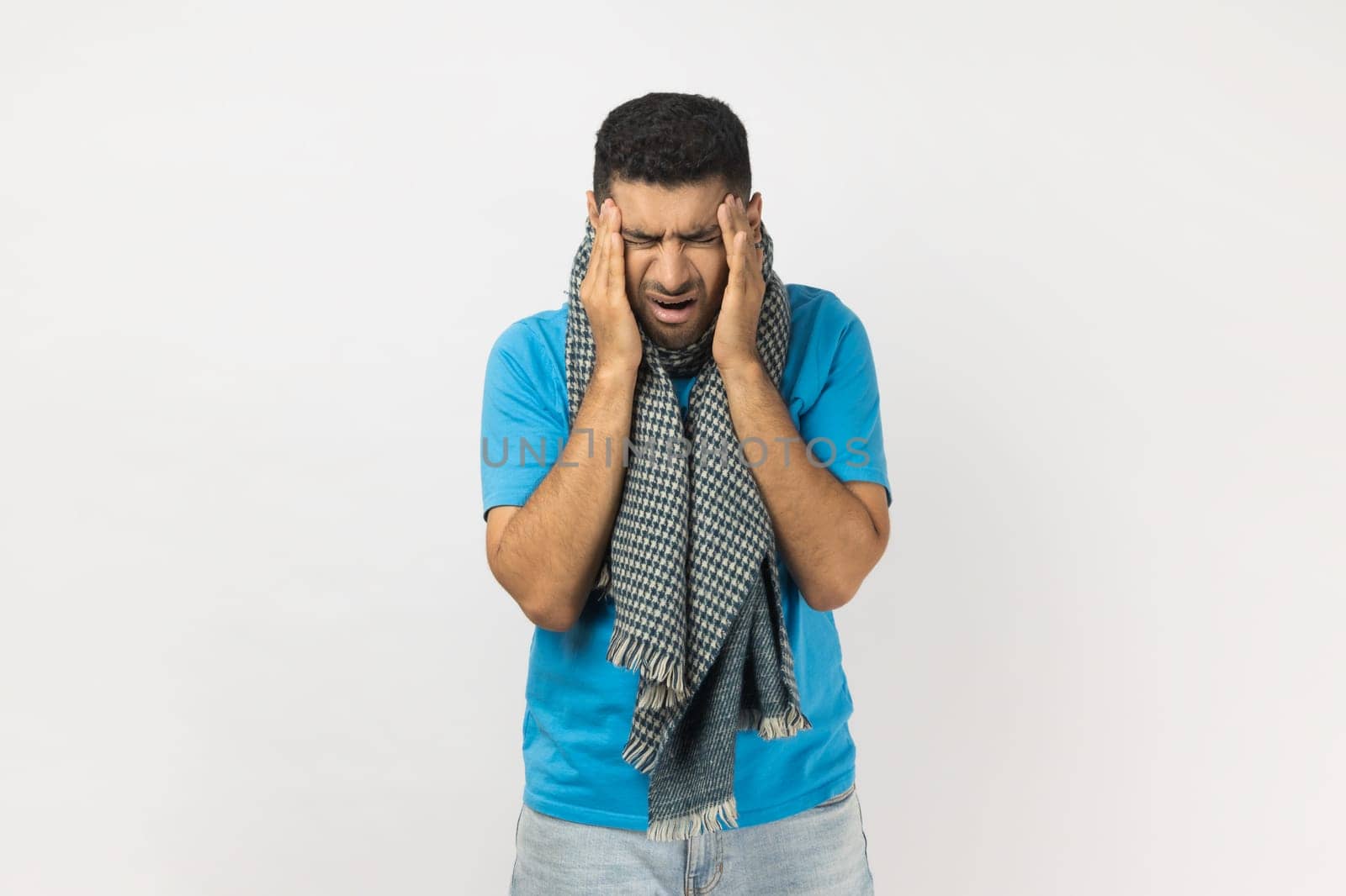 Portrait of sick unshaven man wearing blue T- shirt and wrapped in warm scarf standing, suffering terrible headache, massaging temples. Indoor studio shot isolated on gray background.