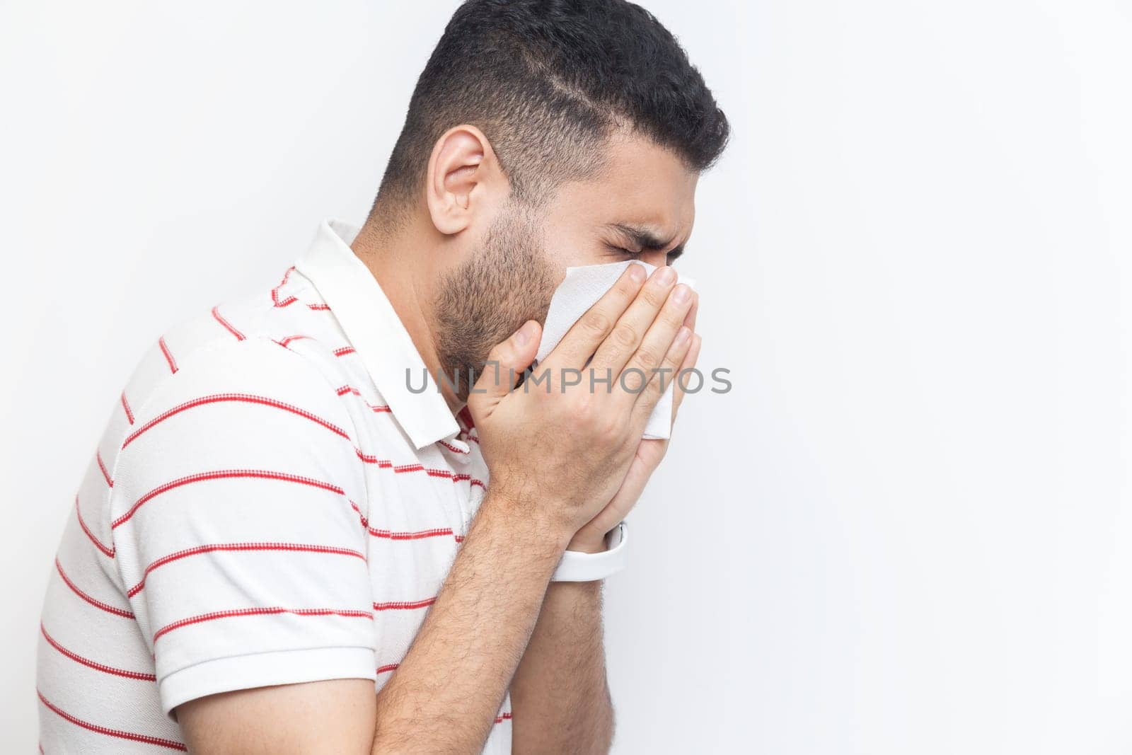 Side portrait of sick unhealthy bearded man wearing striped t-shirt standing having runny nose, using napkin, having flu symptoms. Indoor studio shot isolated on gray background.